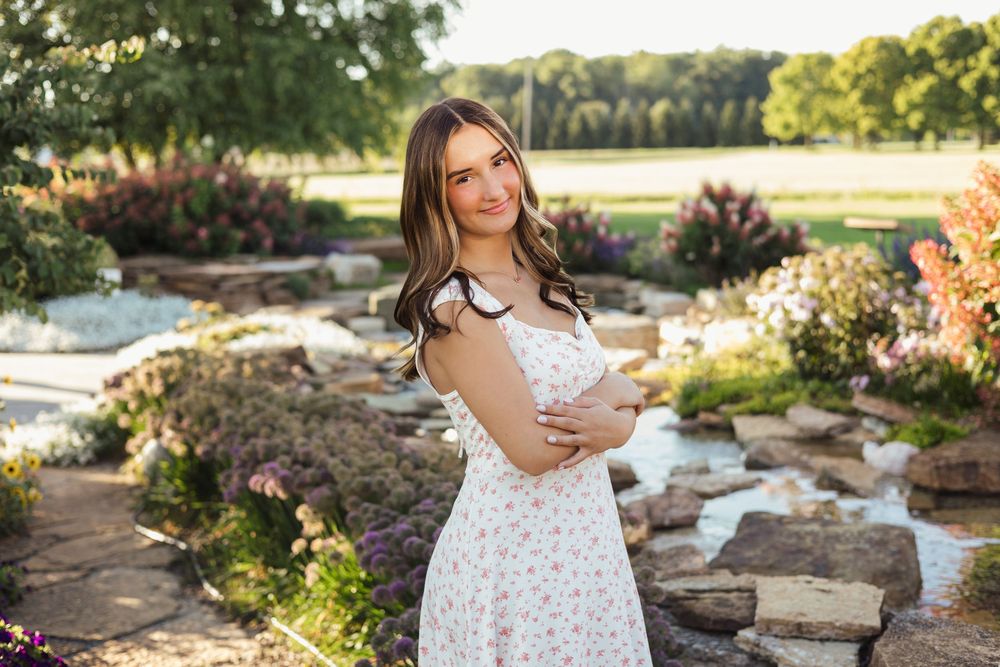Woman in white polka dot dress poses in garden setting with waterfall feature at sunset.