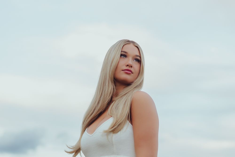 Portrait of a woman with long blonde hair in a white top against a cloudy sky background.