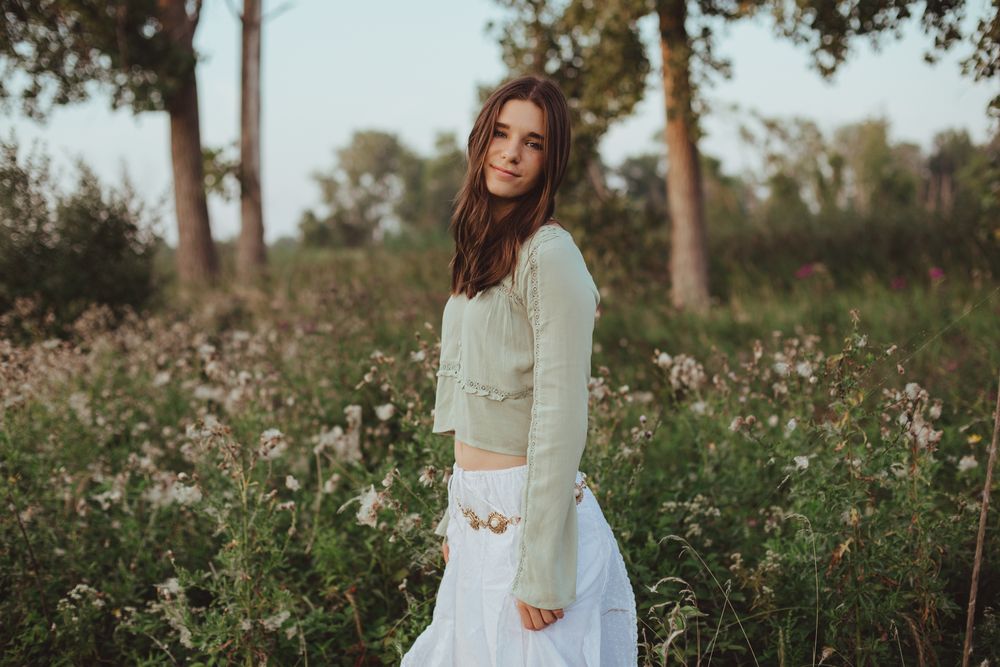 Woman in a light sweater and white skirt poses in a wildflower field at sunset.