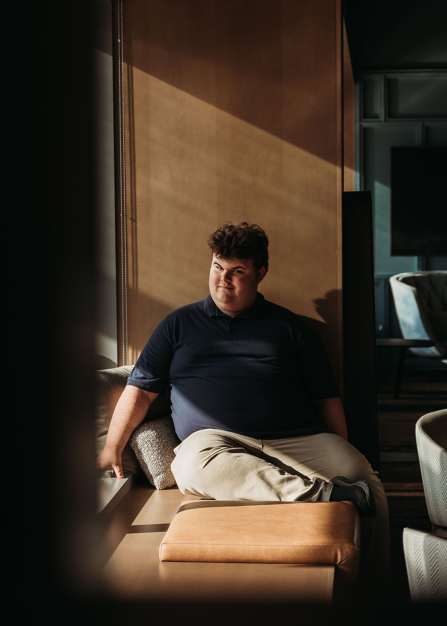 Person relaxing on tan leather couch by window with dramatic lighting and shadows creating moody atmosphere.