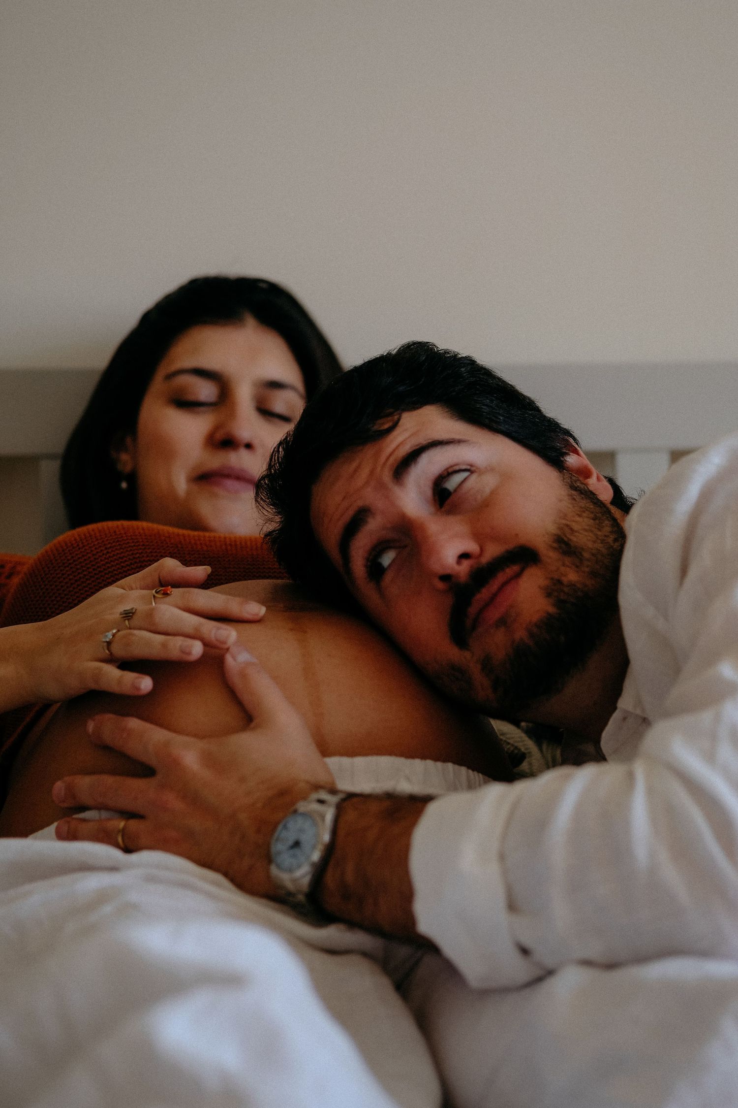 Expecting couple lying in bed together, hands resting on baby bump — intimate maternity moment captured at home.