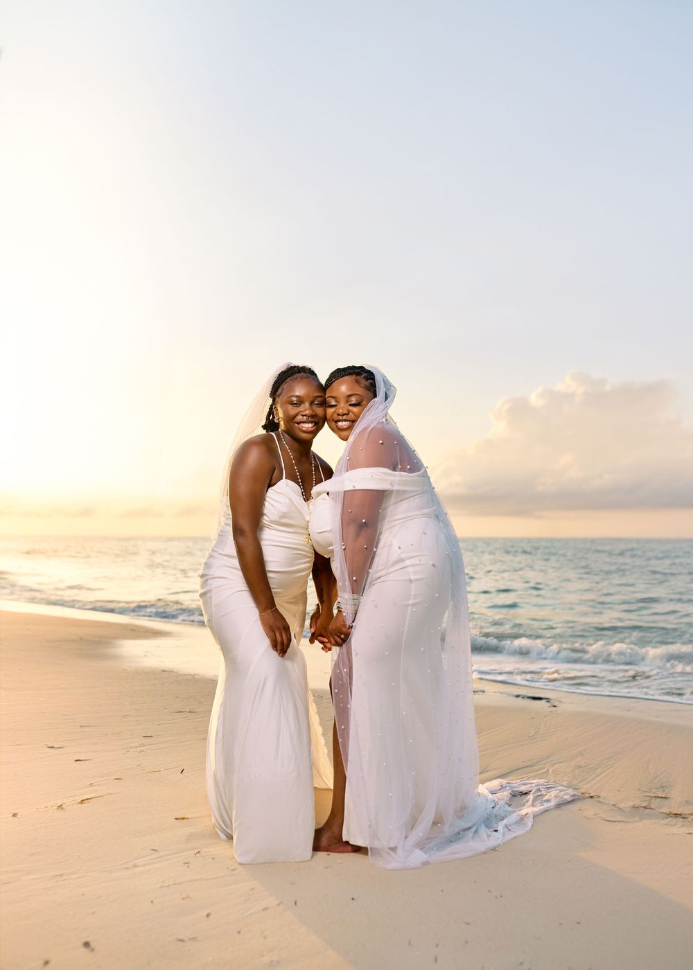 Couple wearing white dresses share an intimate moment on a beach at sunset with ocean waves in the background.