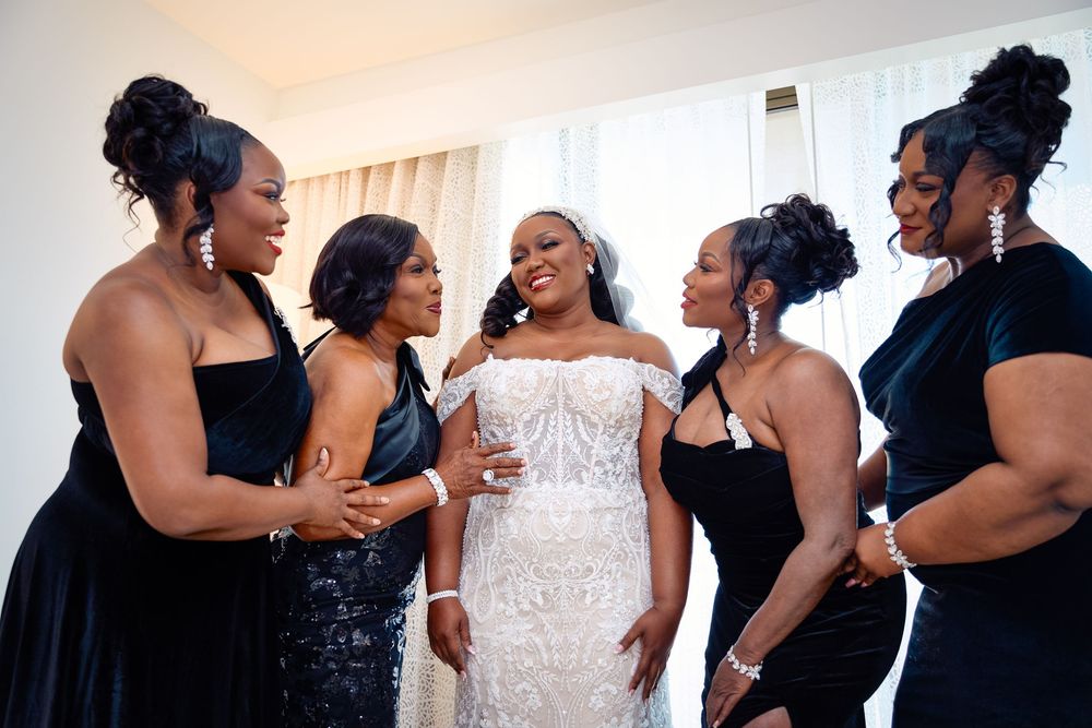 Bride in white lace gown surrounded by bridesmaids in black dresses, smiling together before wedding ceremony.