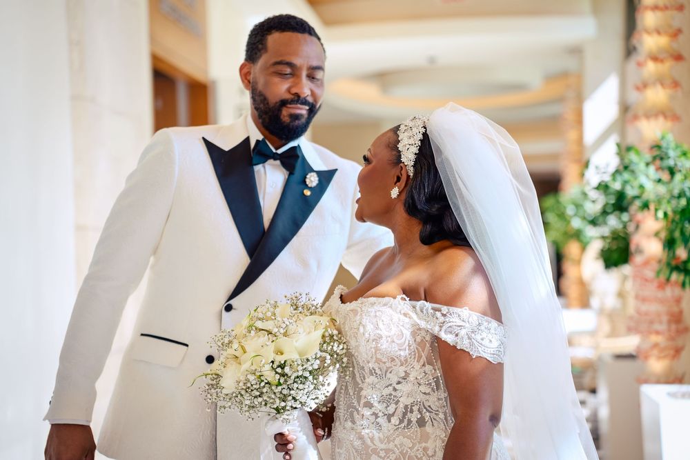 A bride and groom in elegant wedding attire smile at each other, holding a white floral bouquet.