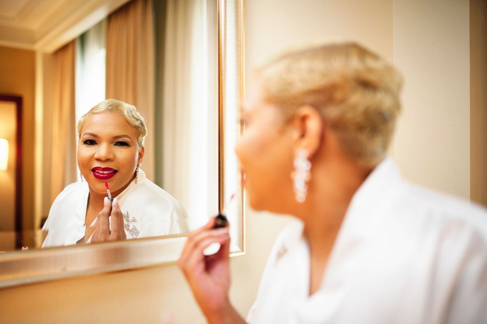 A reflection in a mirror shows someone wearing a white bathrobe and pearl earrings applying lipstick while getting ready.