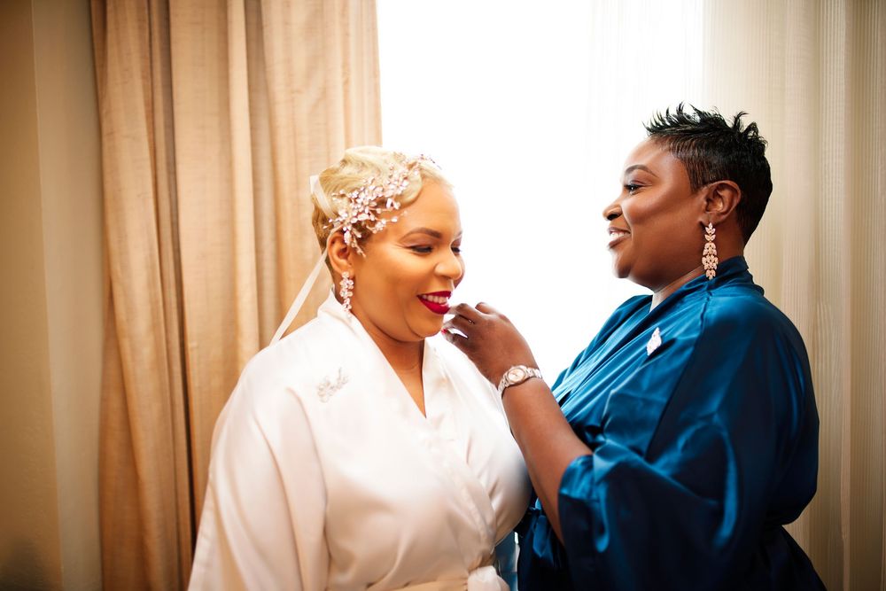 A person in a white robe sits while getting makeup applied by a makeup artist wearing blue.