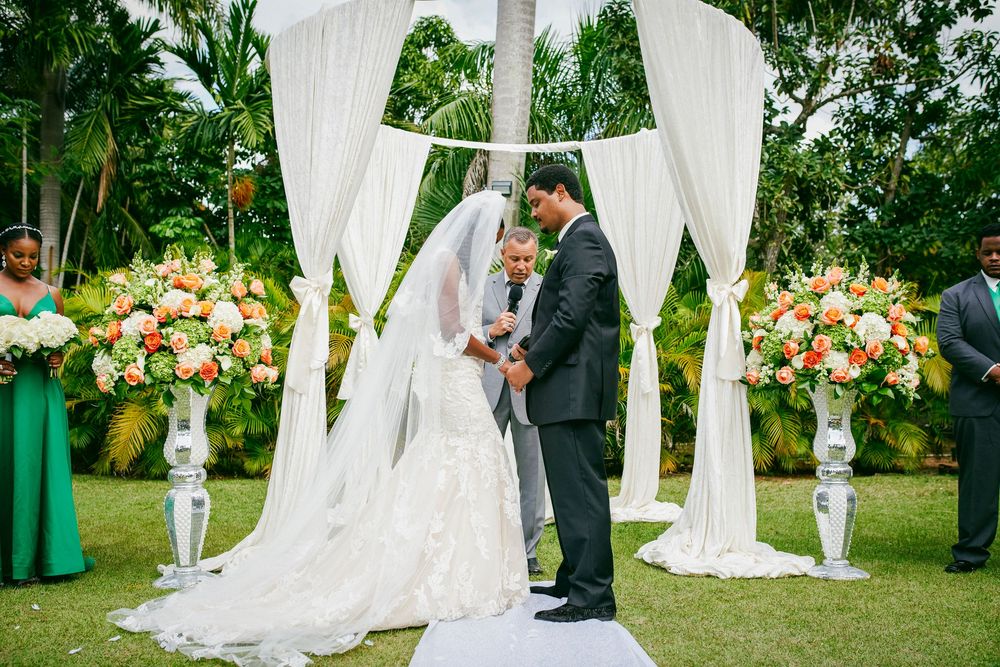 Garden wedding ceremony takes place under an elegant white draped arch with lush floral arrangements and palm trees.