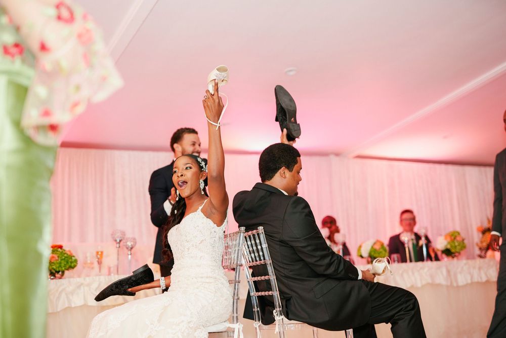 A couple plays a fun wedding shoe game during their reception under pink lighting at a decorated venue.