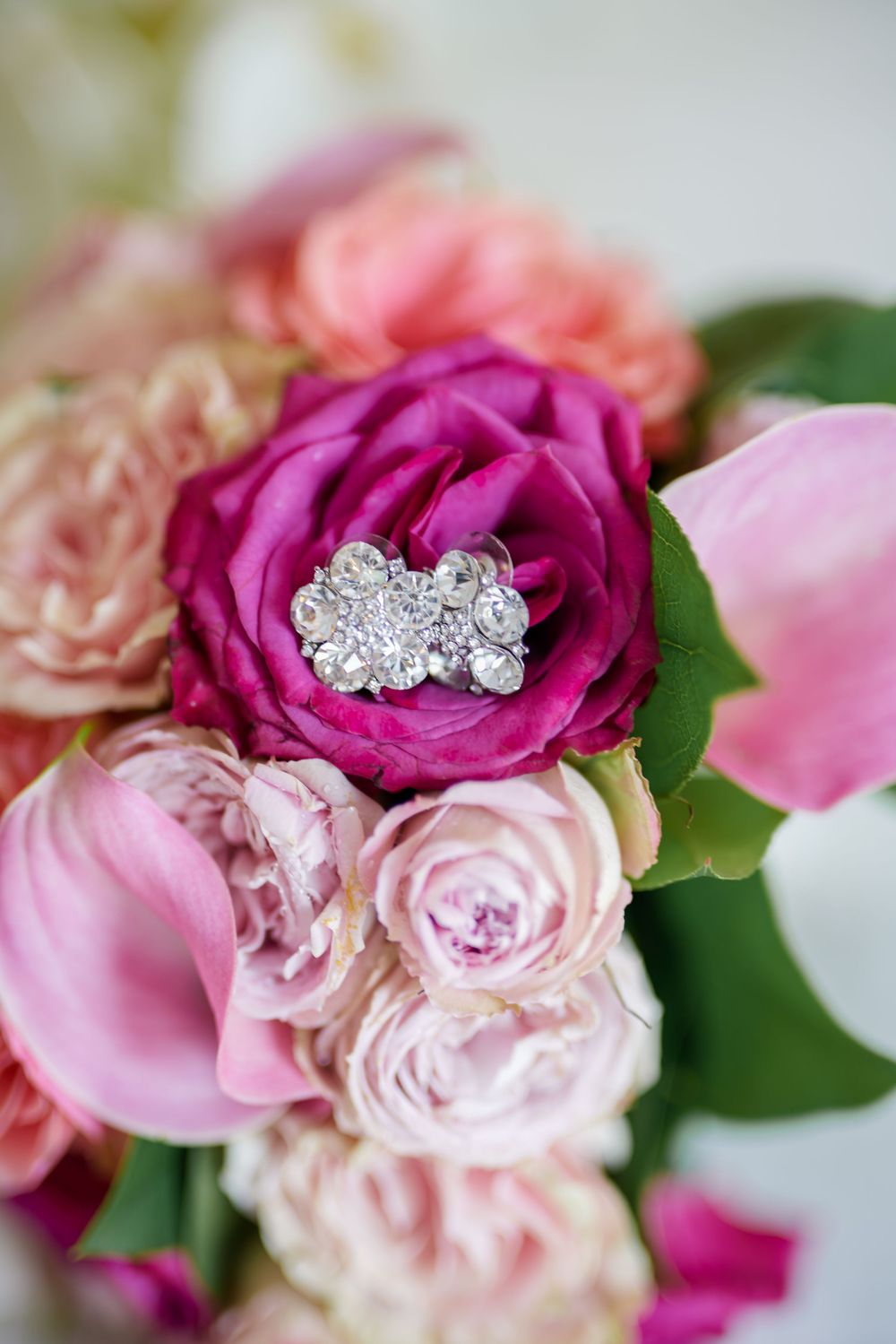 A sparkling diamond engagement ring nestled in pink and magenta roses with green leaves in a bridal bouquet.