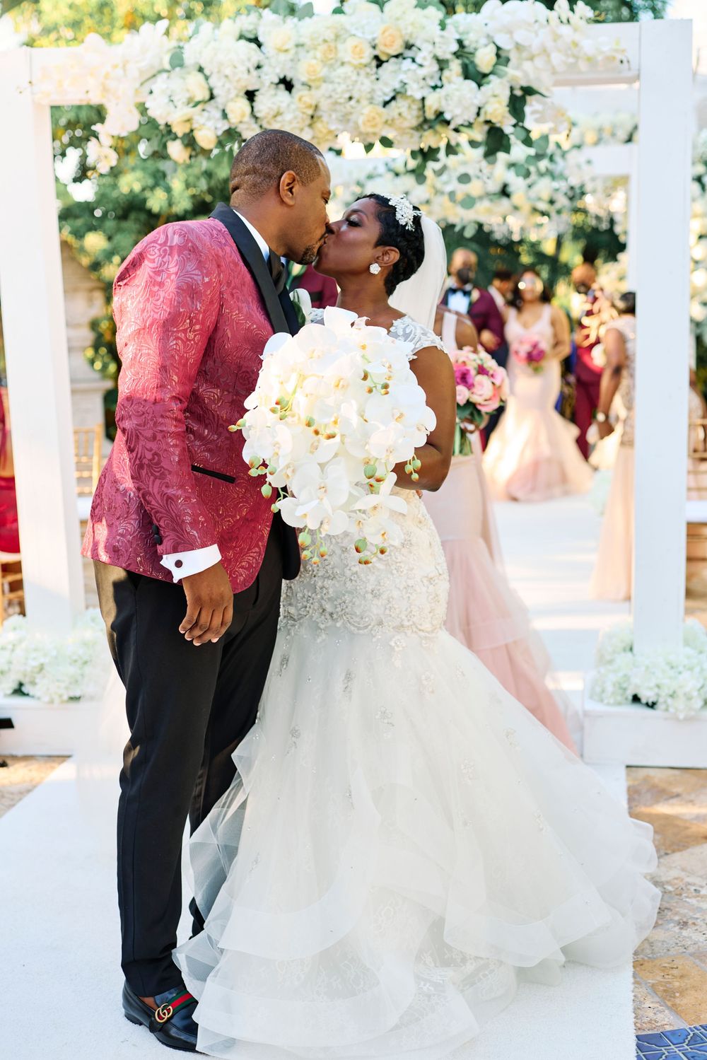 A romantic wedding kiss under a white floral arch, with the groom in a red jacket and bride in a flowing white gown.