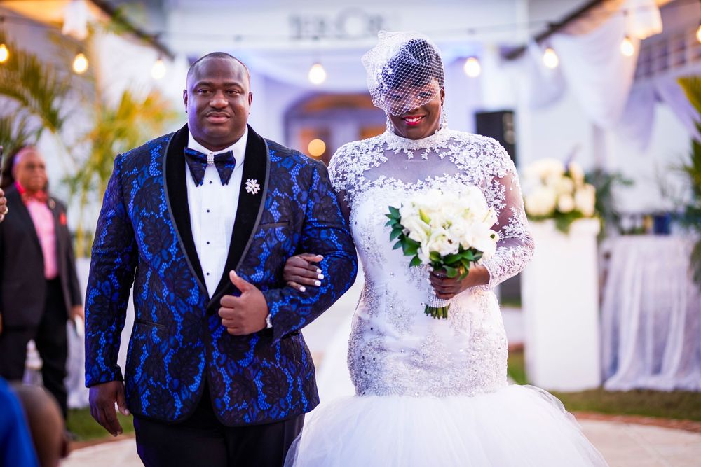 A couple at their wedding ceremony, with the groom wearing a blue patterned tuxedo and the bride in a white lace gown.