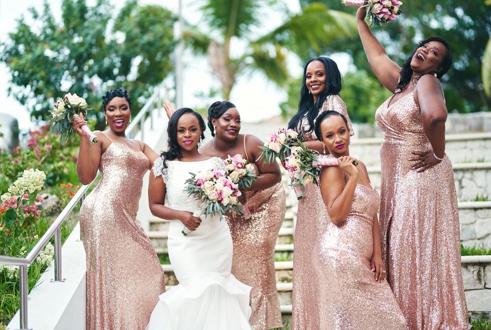 Bridesmaids in sparkly rose gold sequin dresses pose joyfully on outdoor steps with bride in white gown holding bouquet.