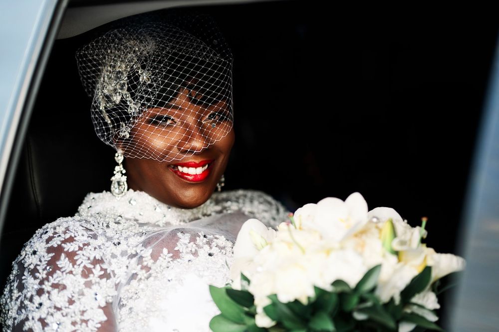A radiant bride in a white lace gown and veil sits in a car with a bouquet of white flowers.