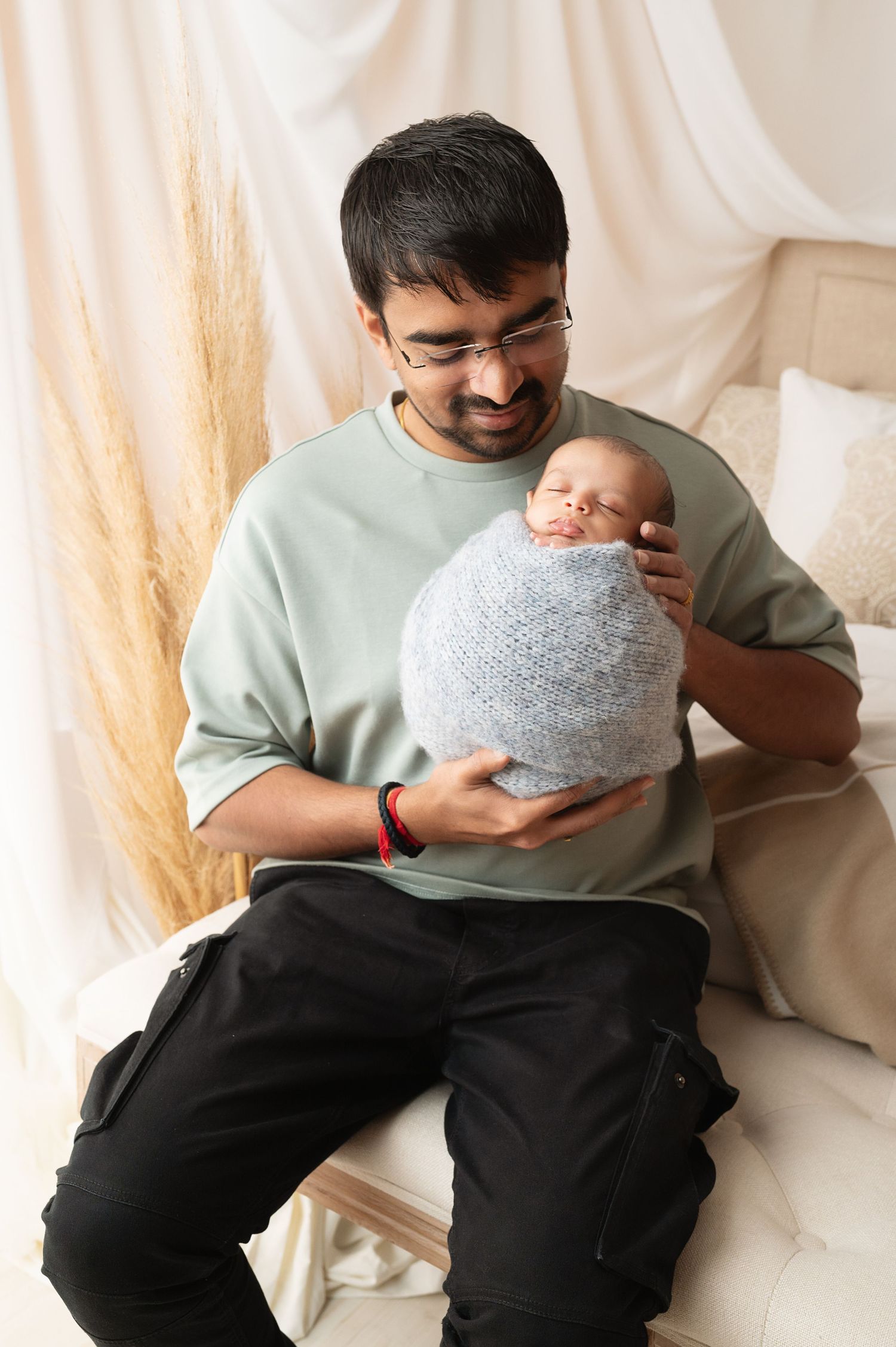 Baby in gray wrap being cradled against mint green sweater with pampas grass decor in background.
