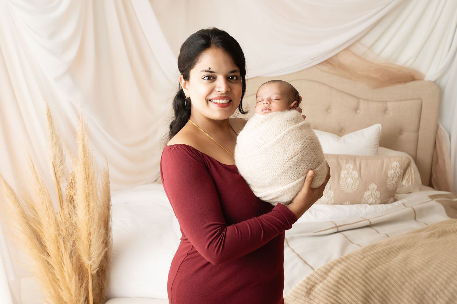 Woman in burgundy dress holding swaddled newborn against white draped backdrop with dried pampas grass.