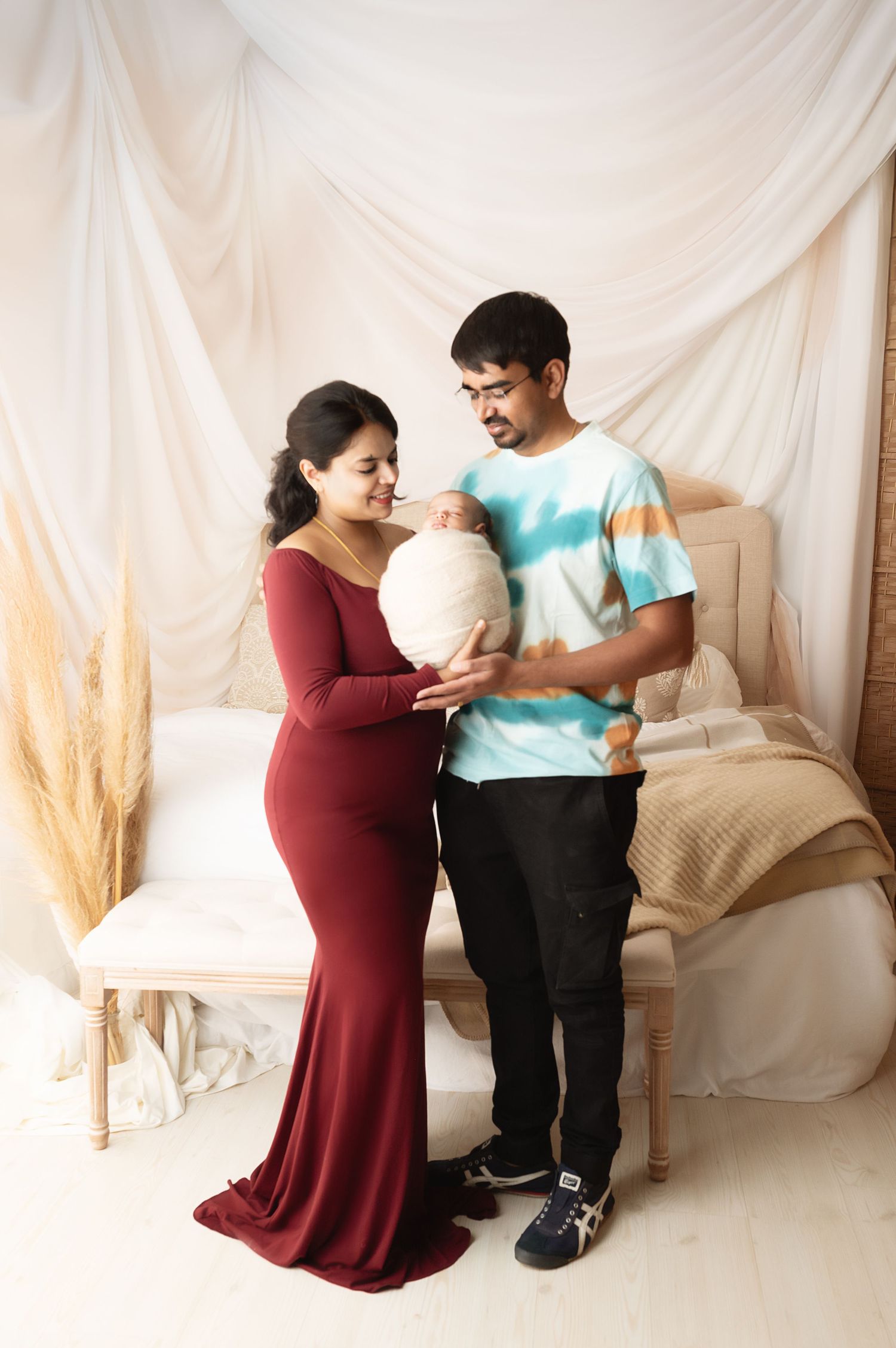 Family portrait with couple in burgundy and teal holding their newborn against draped backdrop.