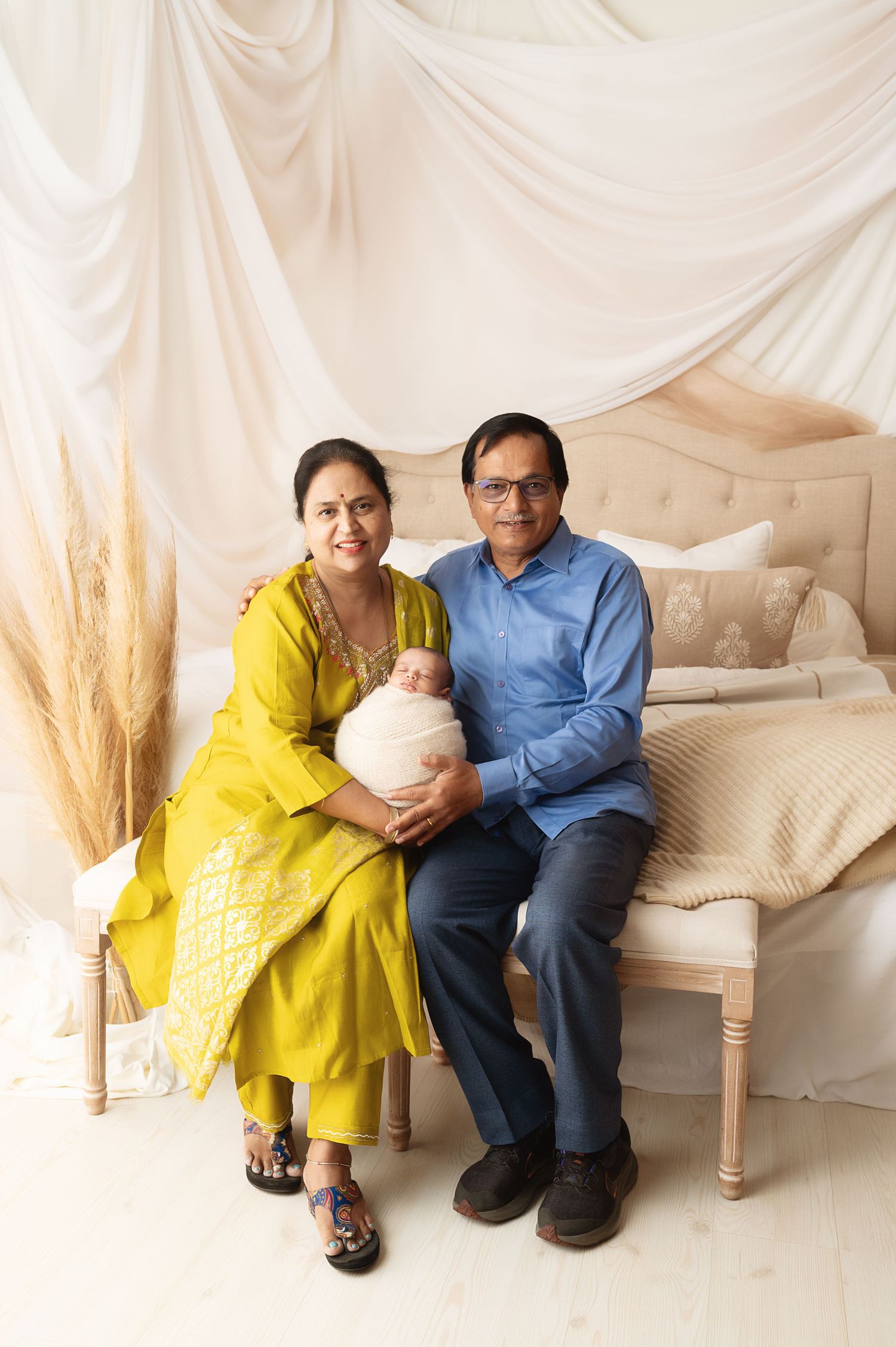 Family portrait with couple in yellow and blue holding newborn on cream bench with pampas grass decor.