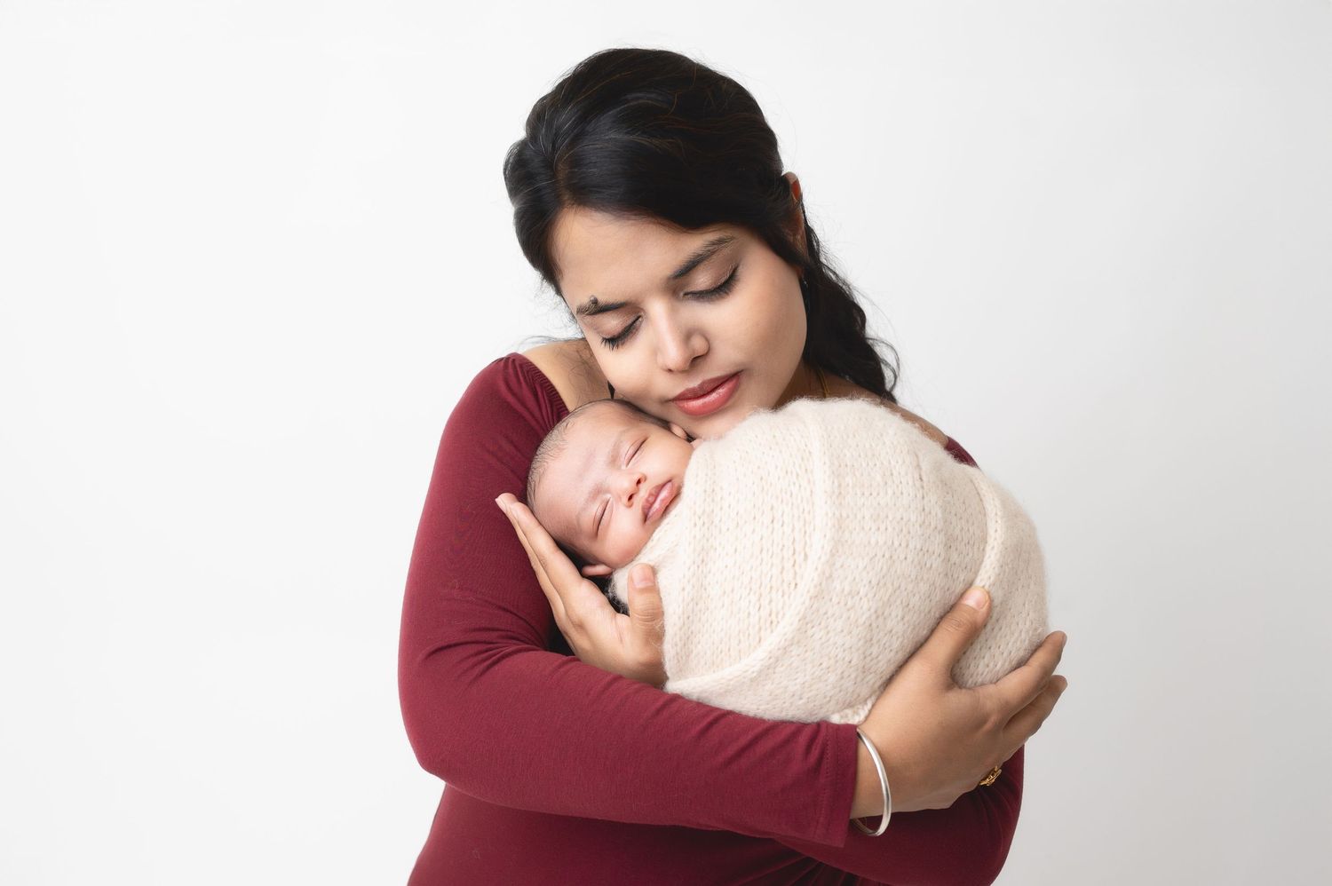 Tender moment between mother in burgundy top and sleeping swaddled newborn against white background.