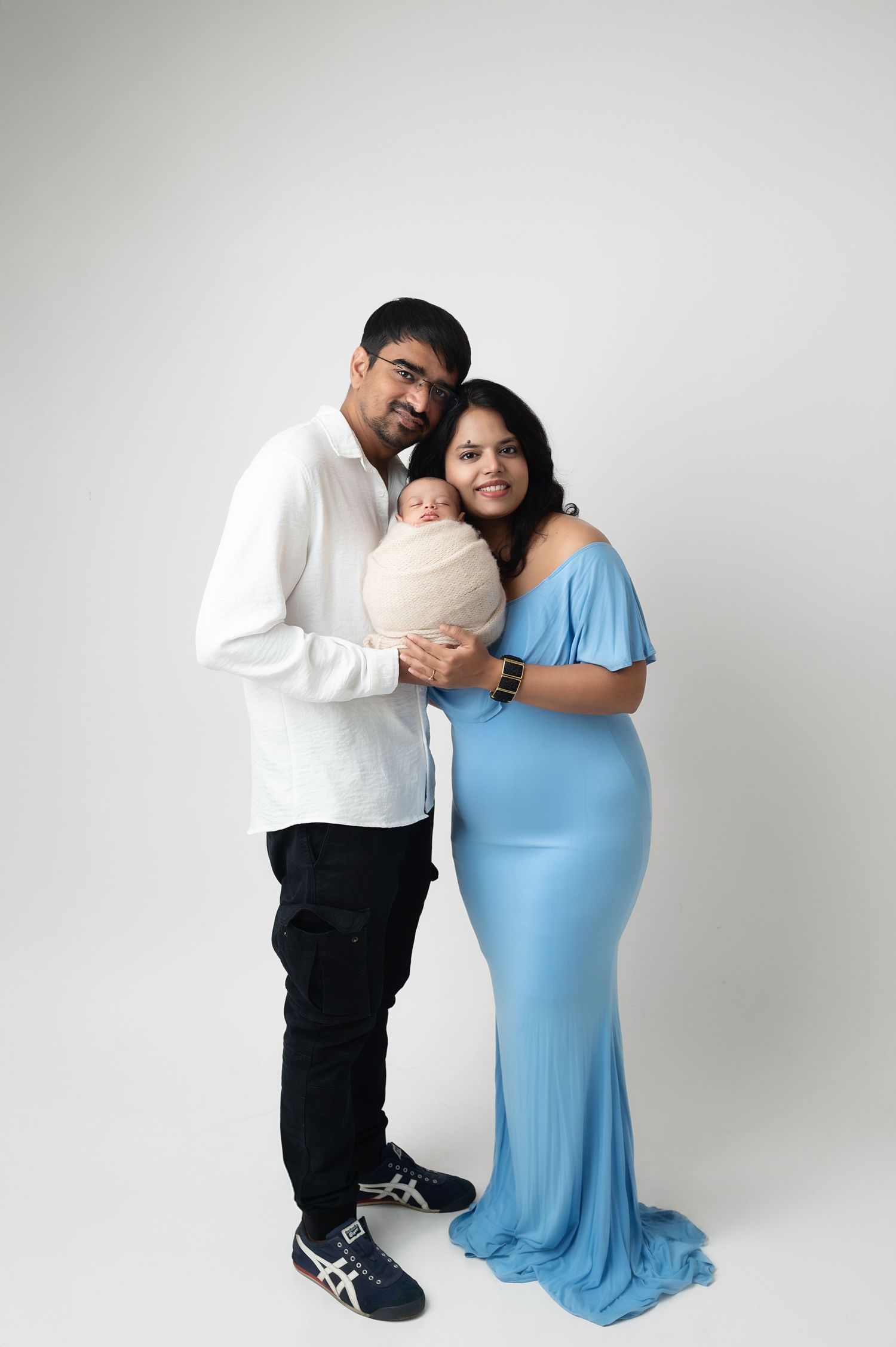Couple in white and blue formal wear holding newborn against minimalist gray backdrop.