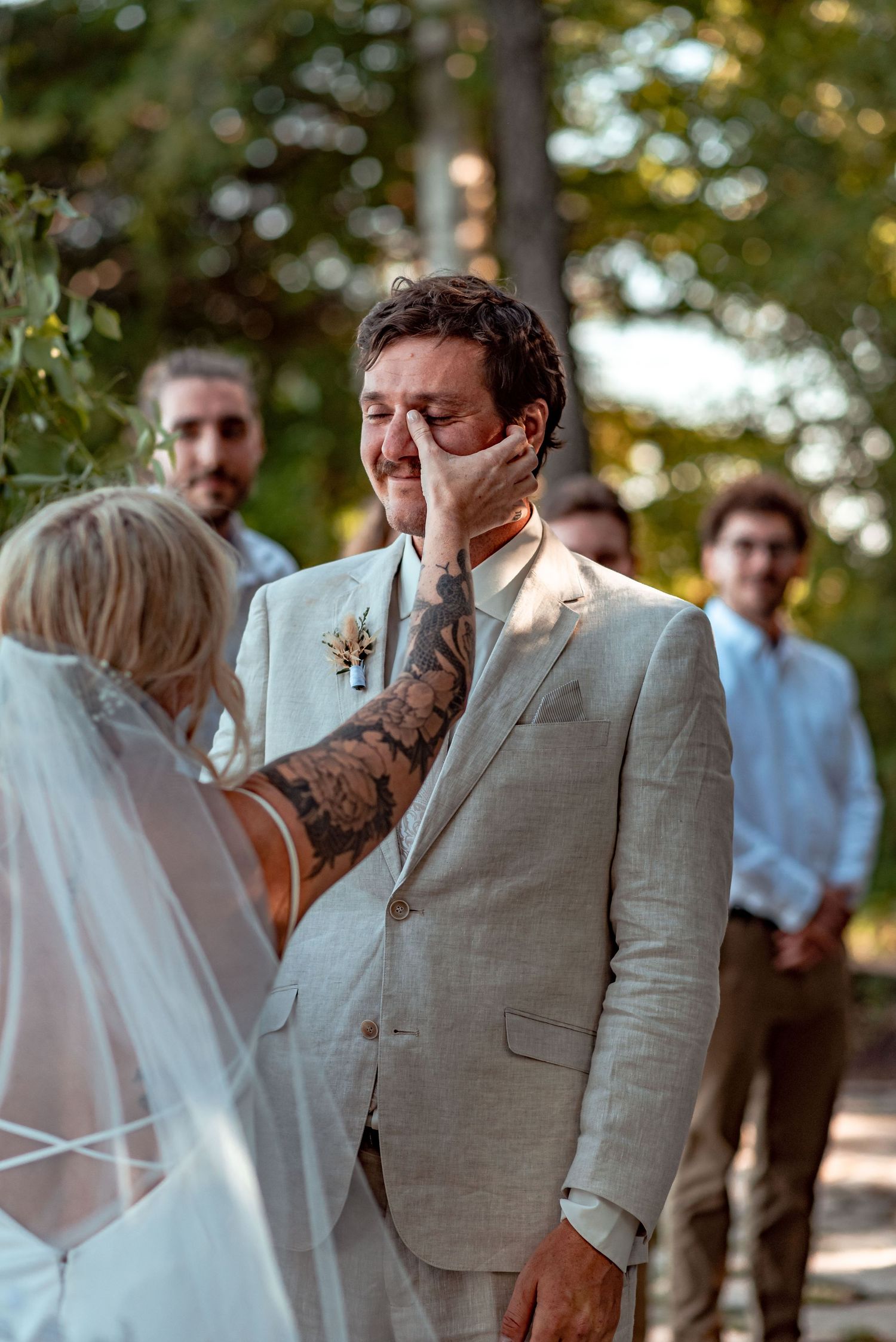 An emotional moment captured during an outdoor wedding ceremony as a groom in a light gray suit appears overcome with feeling.