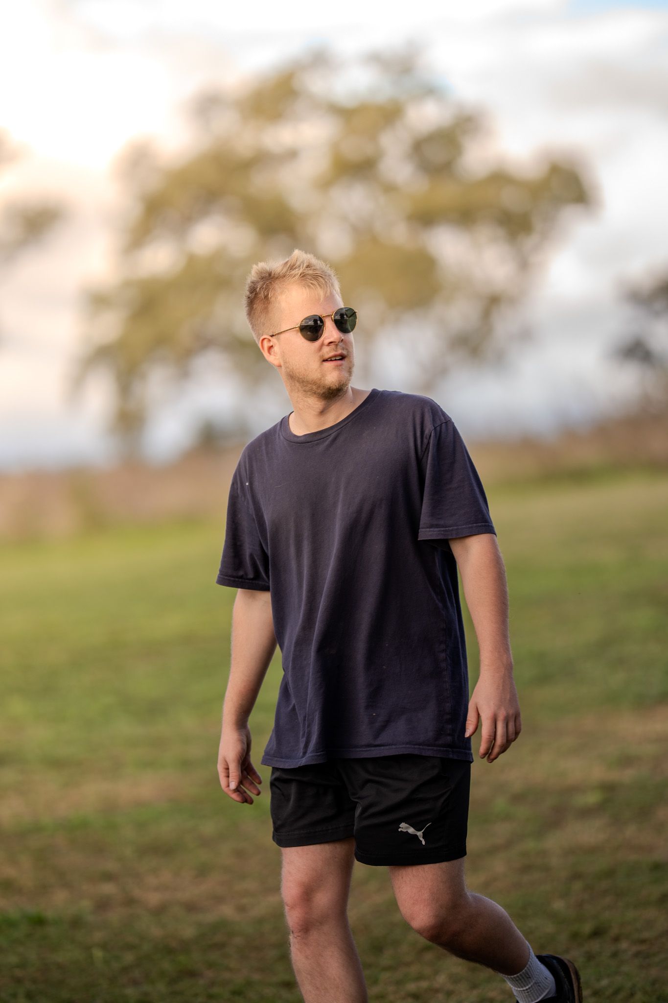 Person wearing sunglasses, dark t-shirt and Puma shorts walking on grassy hill during golden hour.