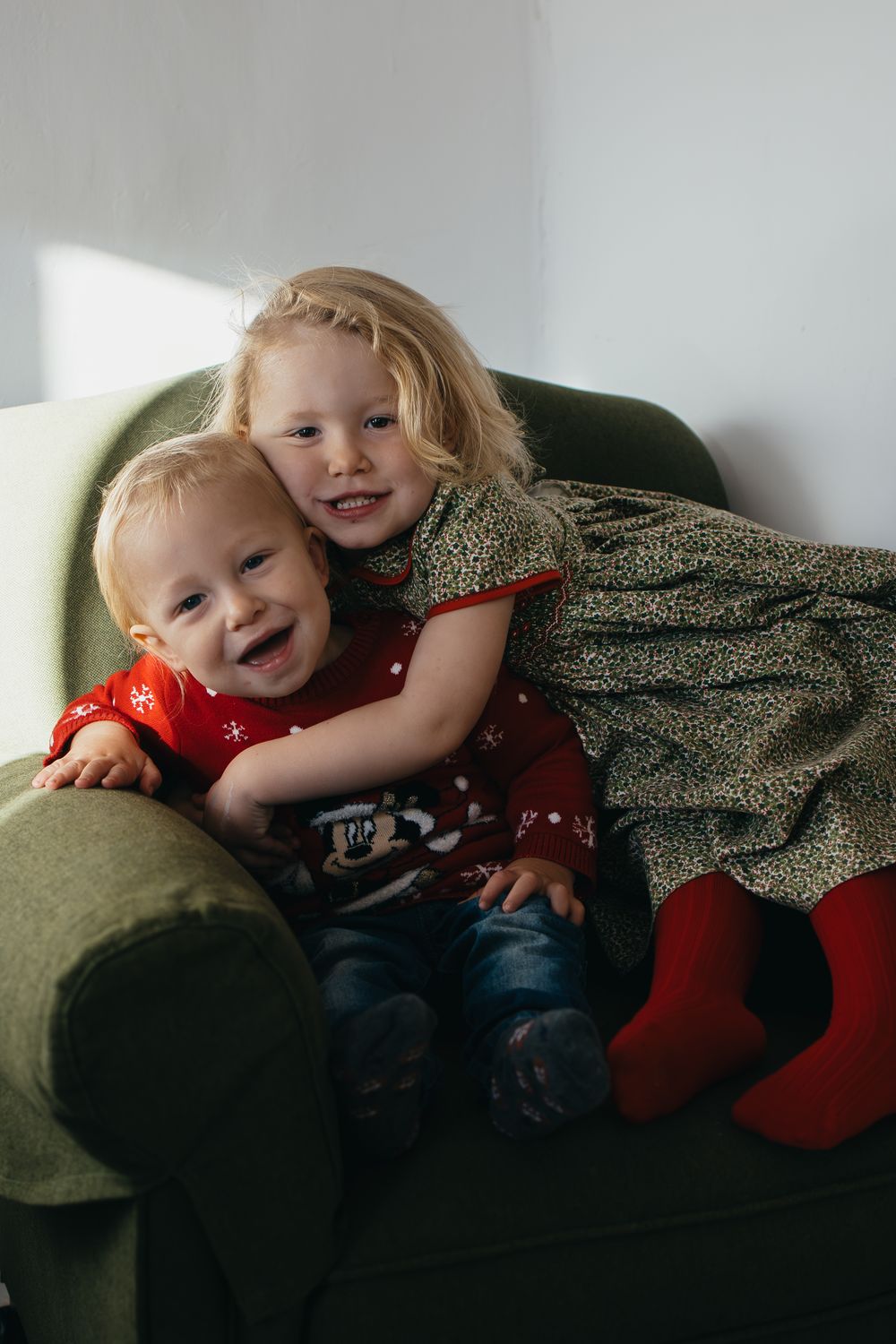 Two young children snuggle together on a green armchair while wearing festive red and green holiday outfits.