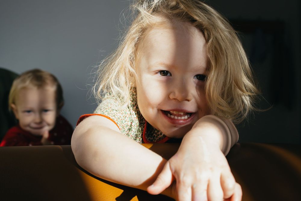 Playful children lean over a dark brown leather couch in natural lighting creating a candid moment.