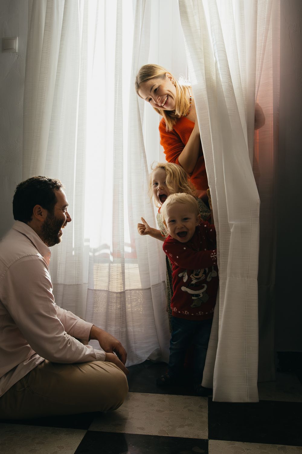 Family playing hide and seek behind white curtains in a sunlit room during a joyful moment at home.