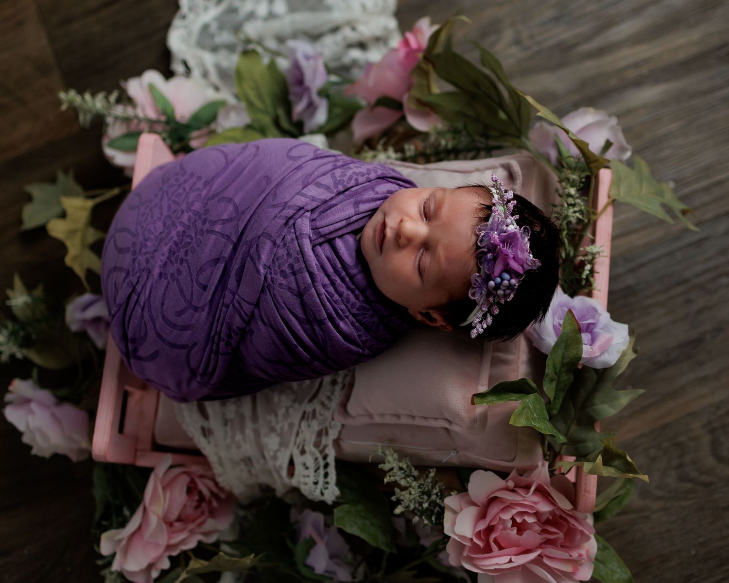 Newborn baby swaddled in purple lace wrap with floral headband, sleeping peacefully in a flower-adorned pink wooden bed.