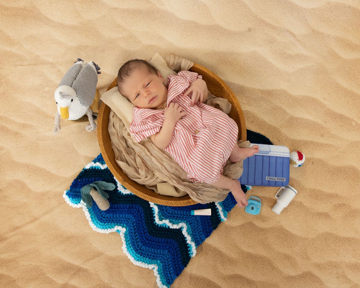 Sleeping baby in pink dress rests in wooden bowl atop blue crochet blanket with toys on sand-textured backdrop.