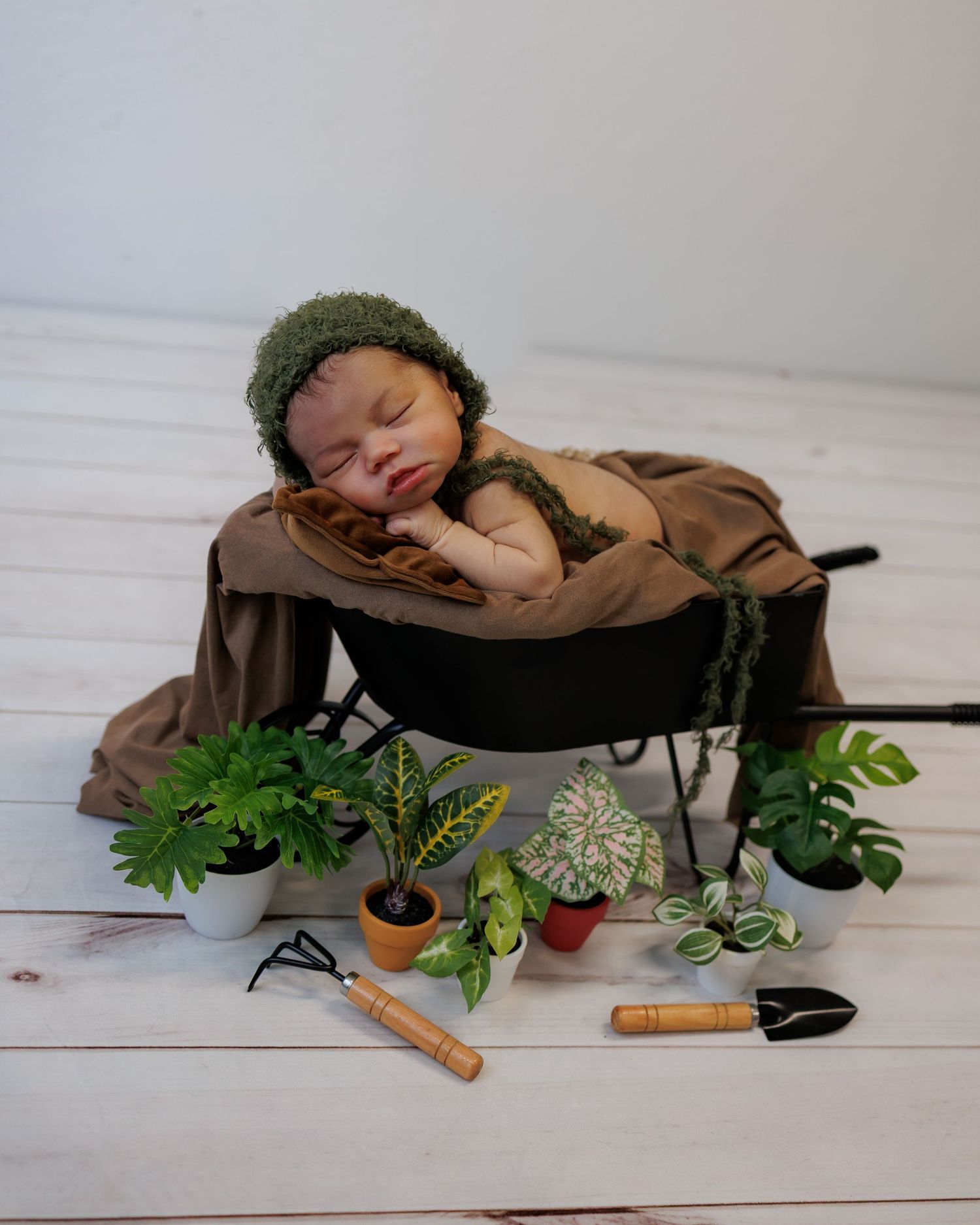 Sleeping newborn baby in green bonnet nestled in a black pot surrounded by miniature garden tools and plants.