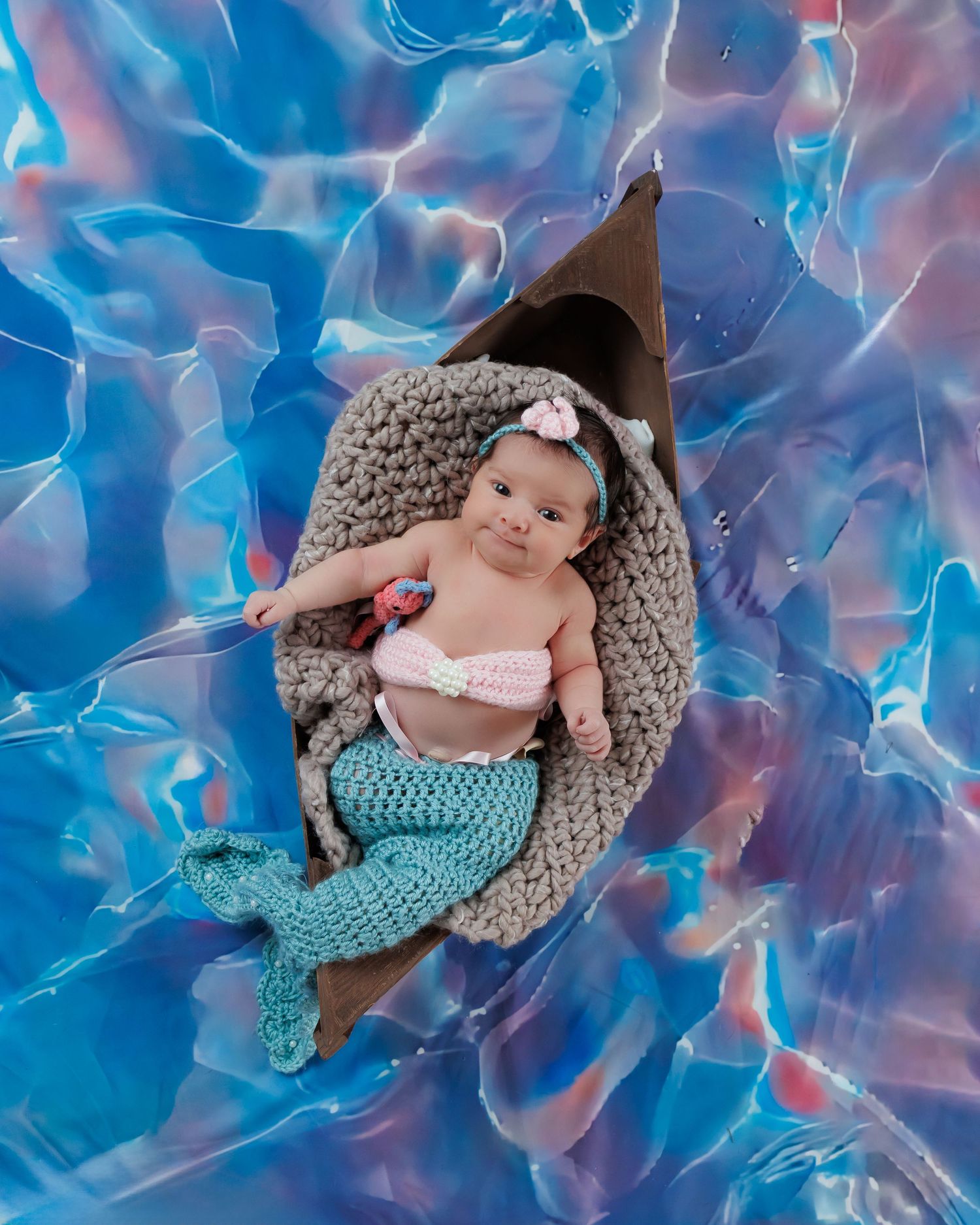 A colorful baby photo featuring a teal mermaid tail costume and gray snug cocoon against a flowing blue backdrop.
