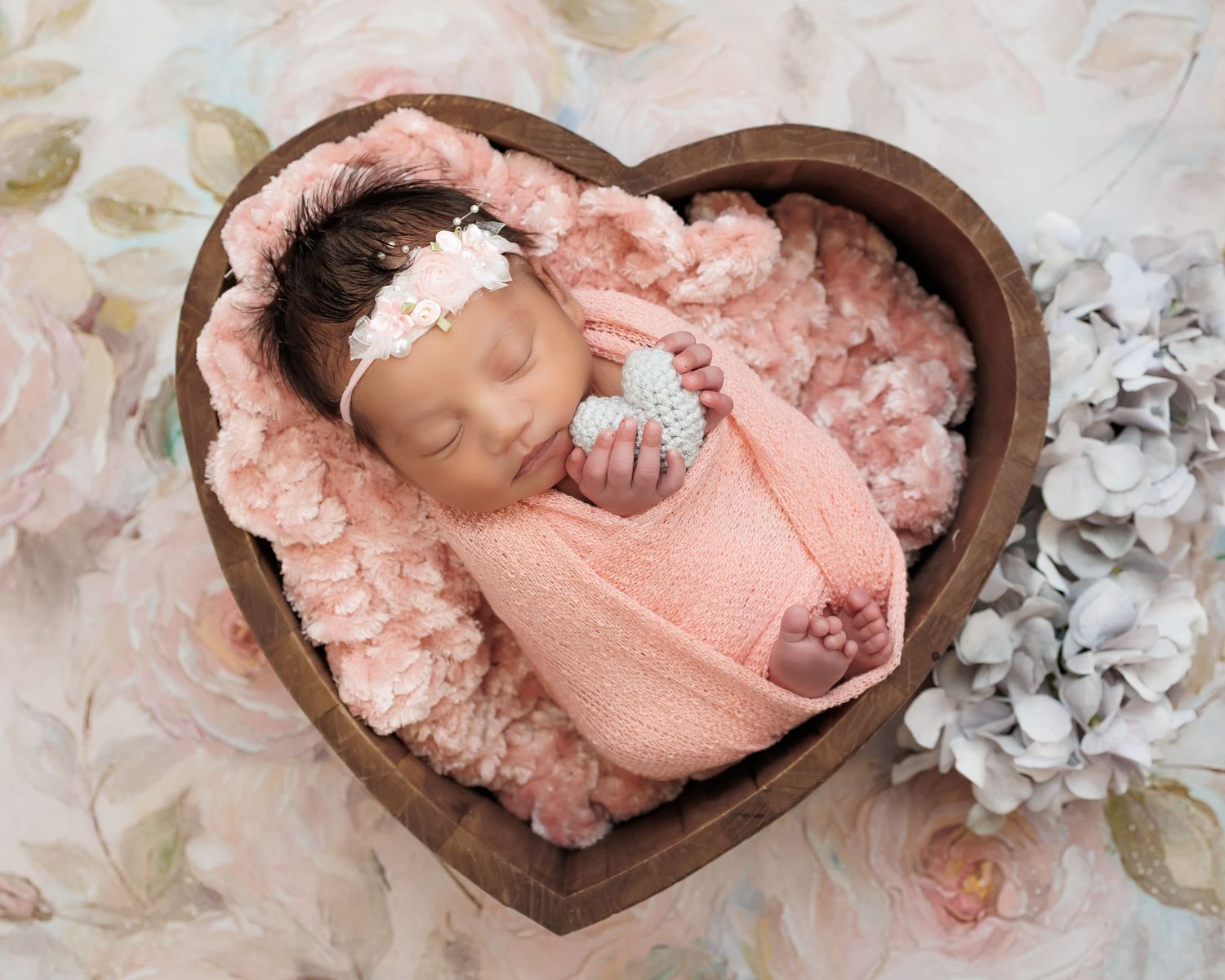 Sleeping newborn wrapped in soft pink swaddle lying in heart-shaped wooden bowl surrounded by delicate white flowers.
