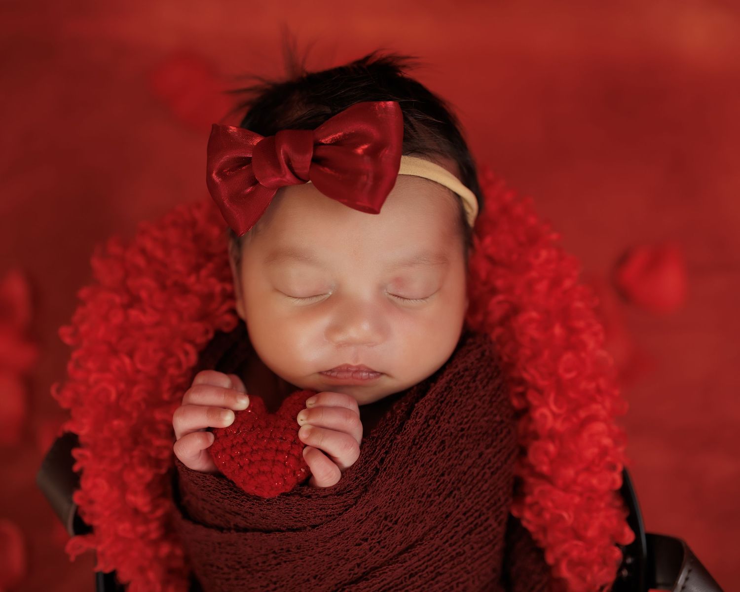 Sleeping baby wrapped in burgundy swaddle with red bow headband on plush red backdrop holding heart decoration.
