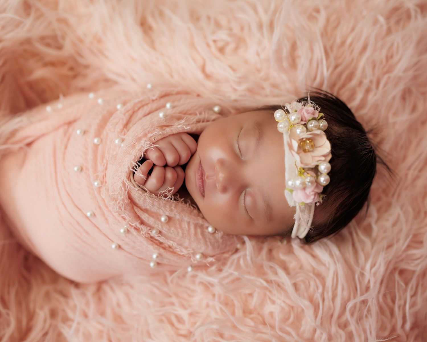 Sleeping newborn baby wearing pink wrap and floral headband rests peacefully on soft peach-colored fur blanket.