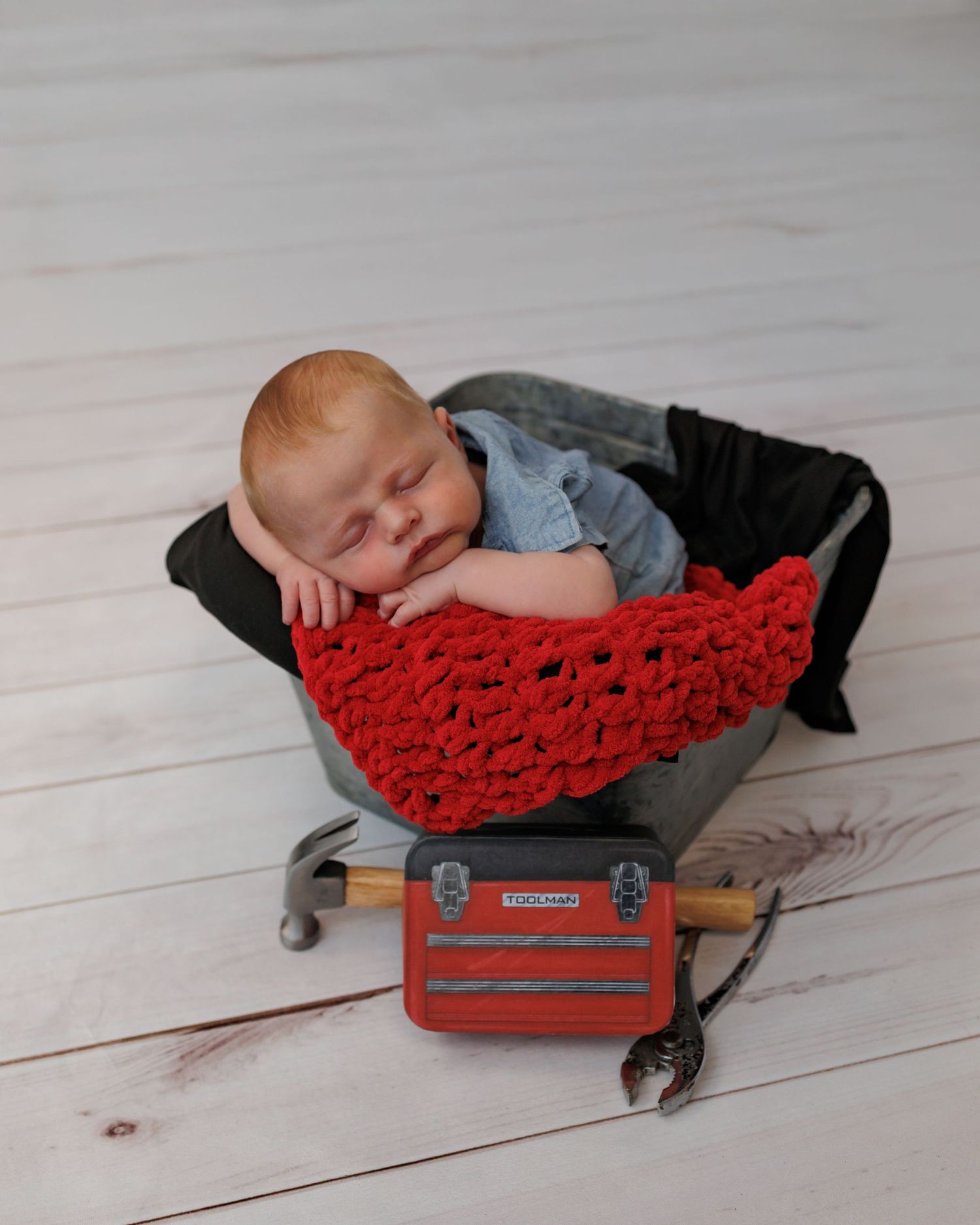 A sleeping baby lies on a red knit blanket atop a vintage red suitcase on a white wooden floor.