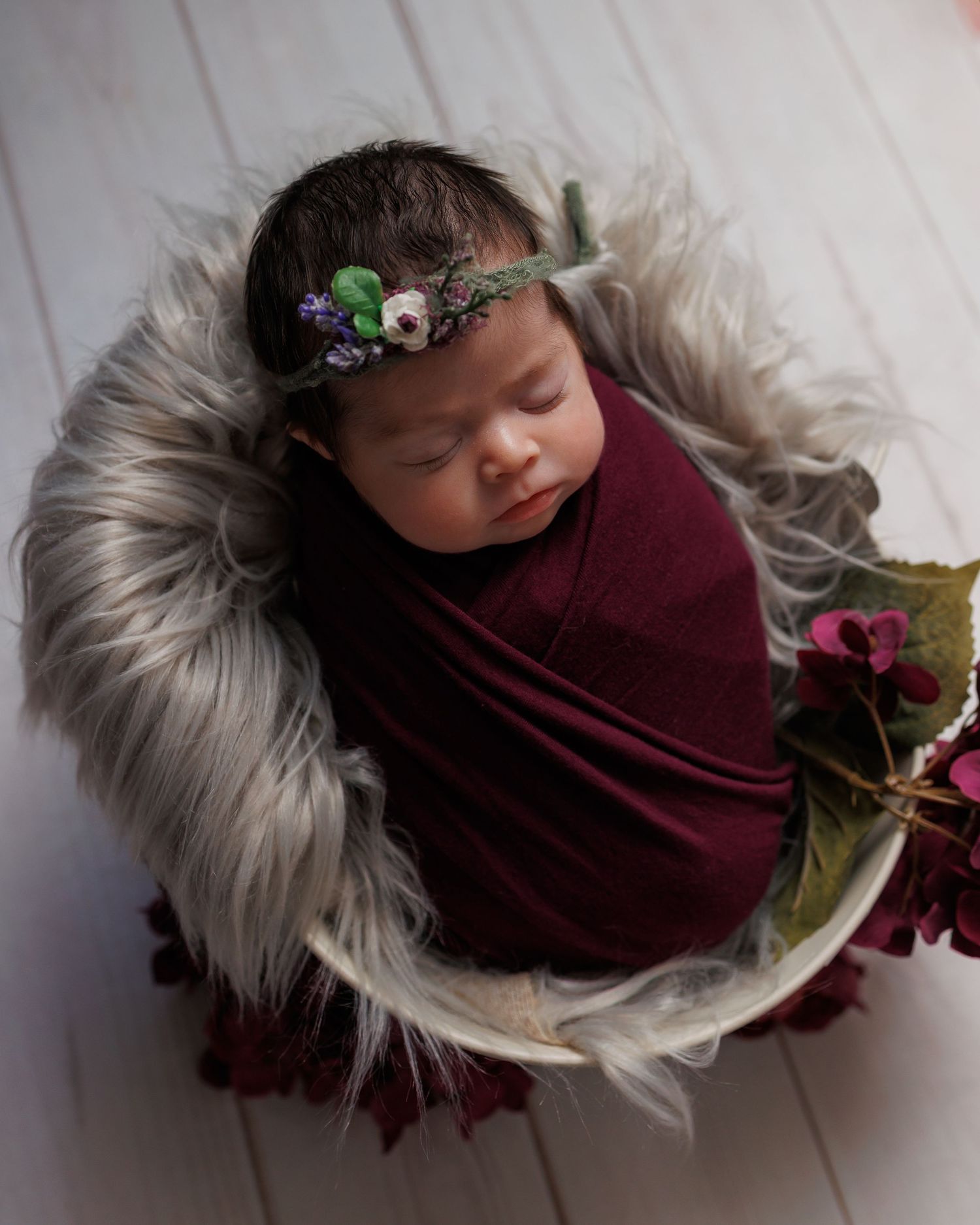 A baby nestled in a plush white fur blanket wearing a burgundy wrap and floral headpiece in a series of angles.