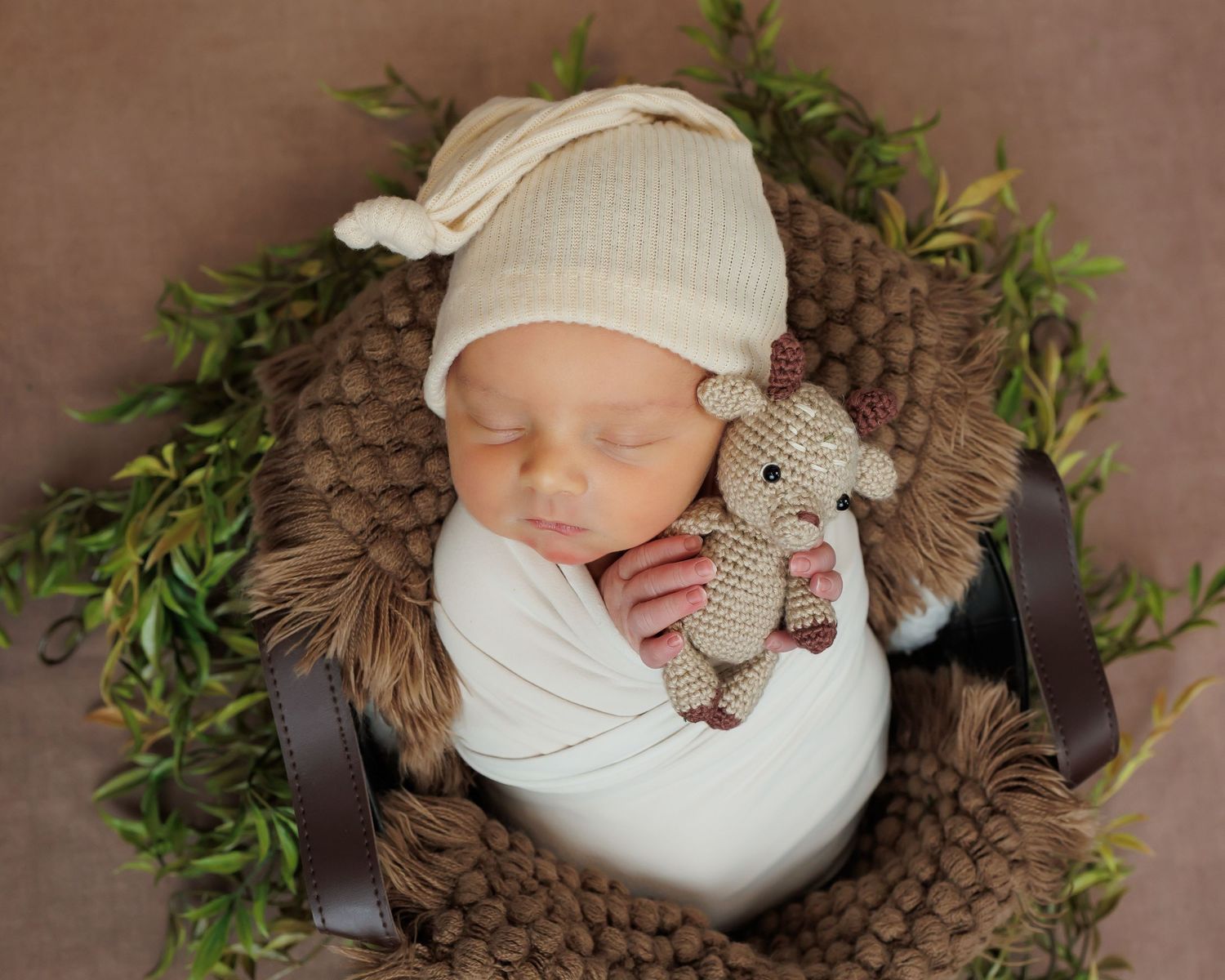 Sleeping newborn wrapped in white swaddle with knit hat and stuffed animal on brown textured blanket surrounded by greenery.
