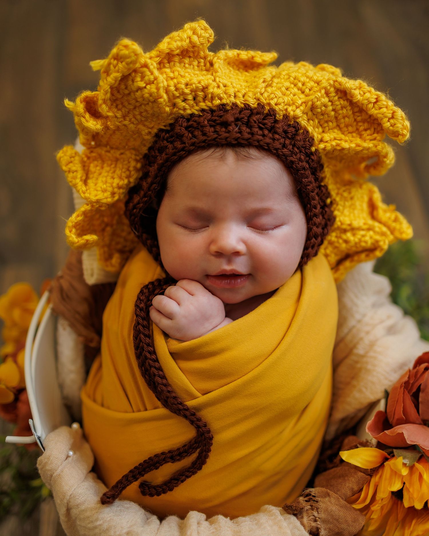 Newborn baby wrapped in mustard yellow swaddle wearing a crochet sunflower bonnet, sleeping peacefully in a small bucket.