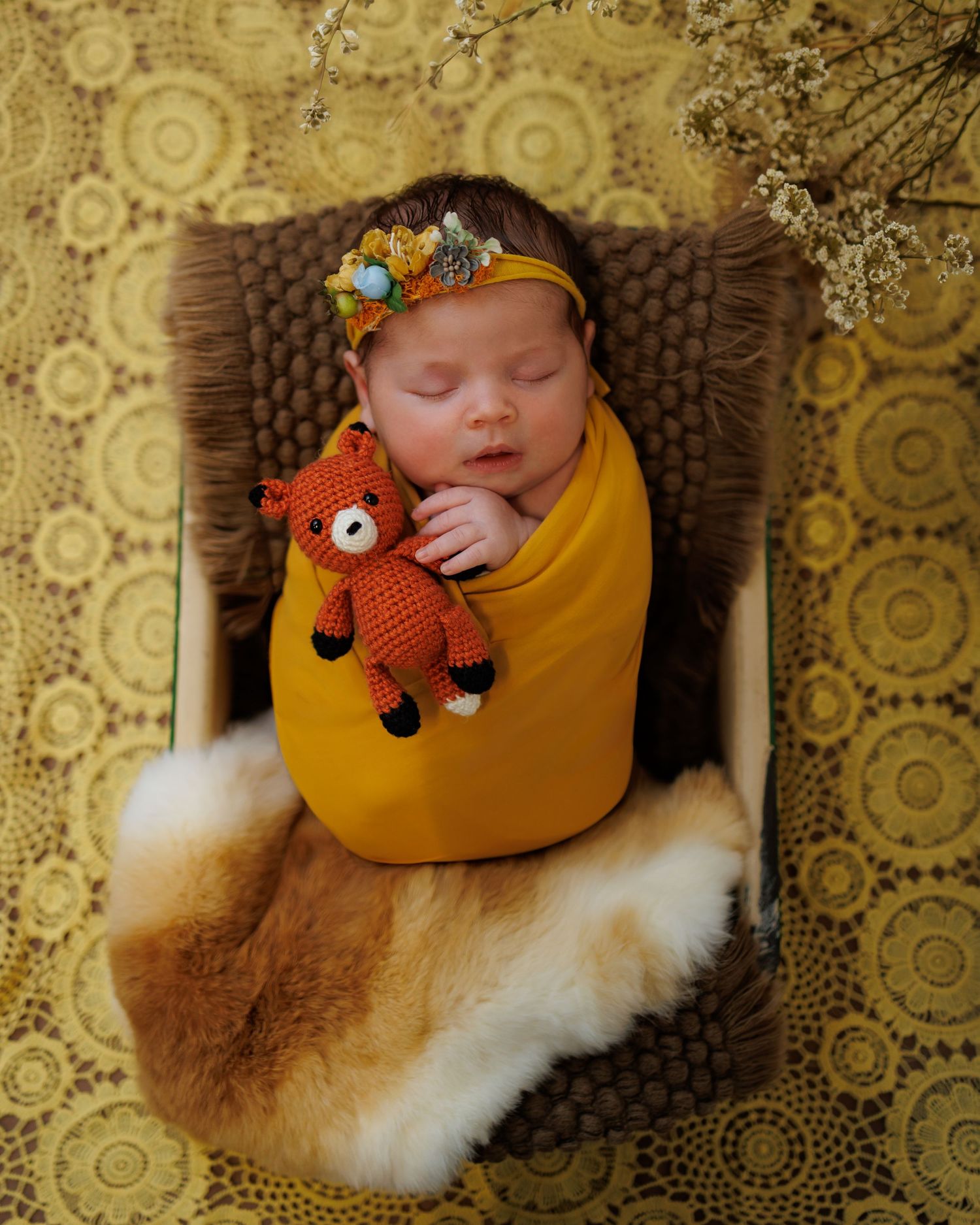 Newborn baby swaddled in mustard yellow wrap, holding a crochet fox toy, wearing a floral crown, sleeping peacefully.