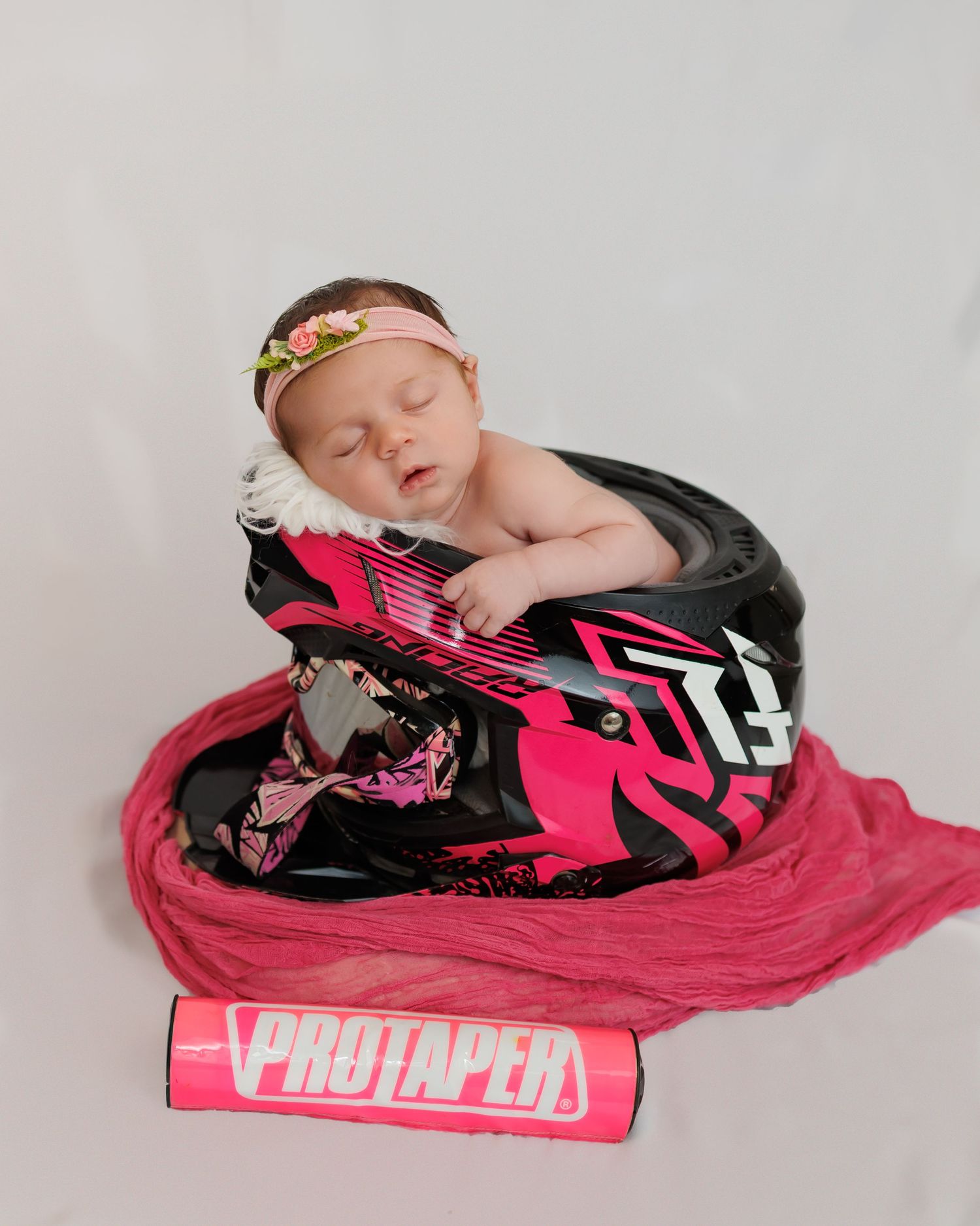 Sleeping newborn wearing floral headband rests peacefully on pink motocross gear and Pro Taper handlebars.