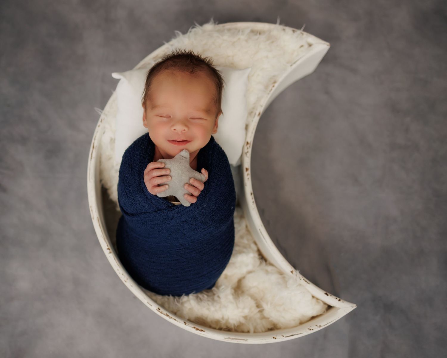 Sleeping newborn wrapped in navy blue swaddle resting in decorative crescent moon prop against gray backdrop.