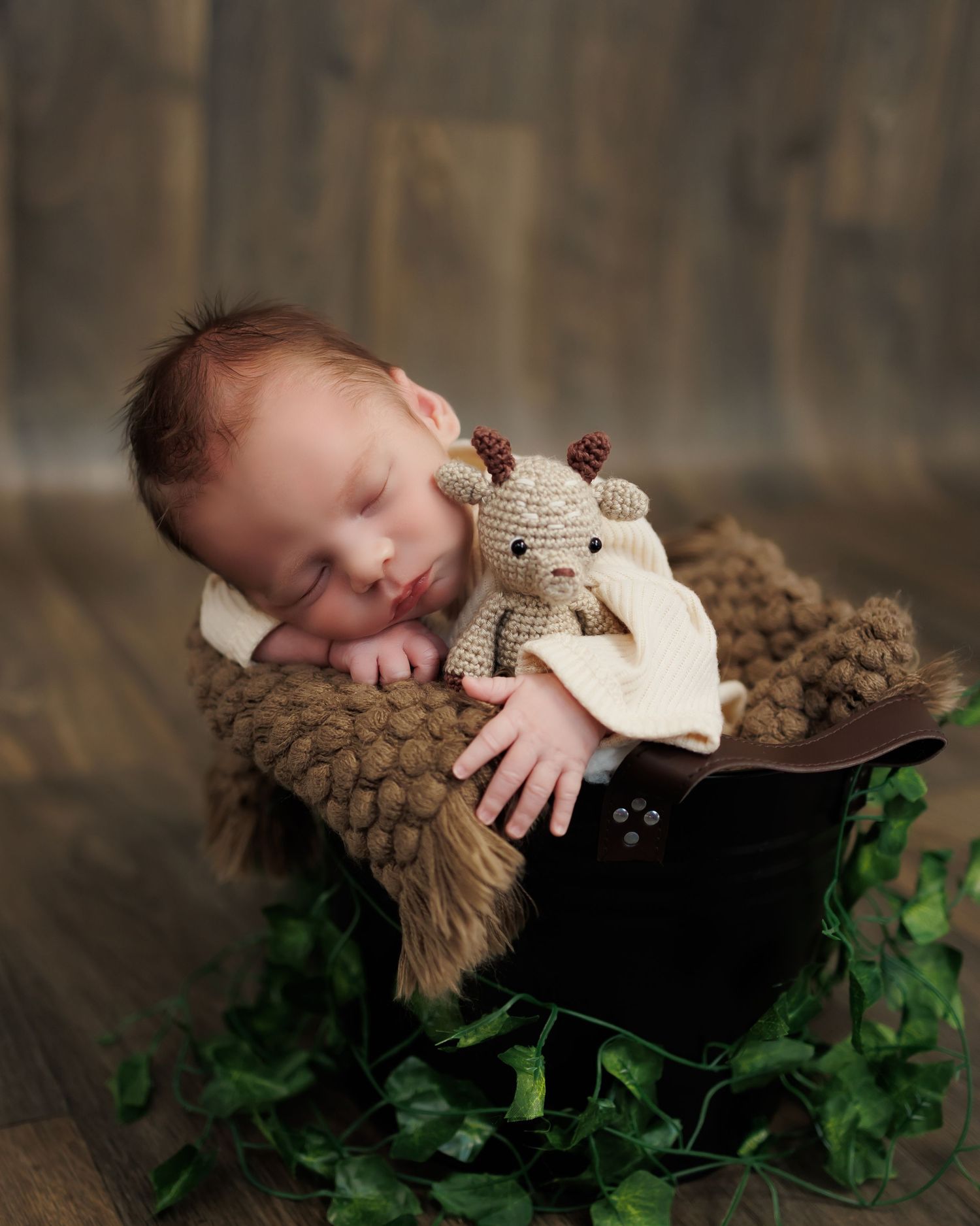 A sleeping newborn rests peacefully in a wooden bowl with a plush teddy bear on a cozy furry blanket surrounded by ivy leaves.