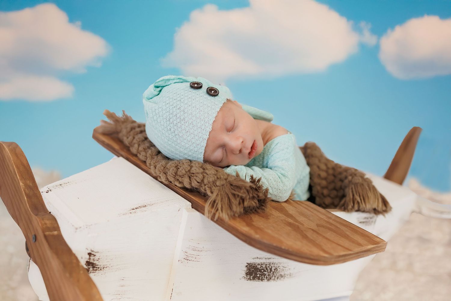 A newborn wearing a light blue knit aviation outfit sleeps on wooden airplane props in a cloud-themed setting.