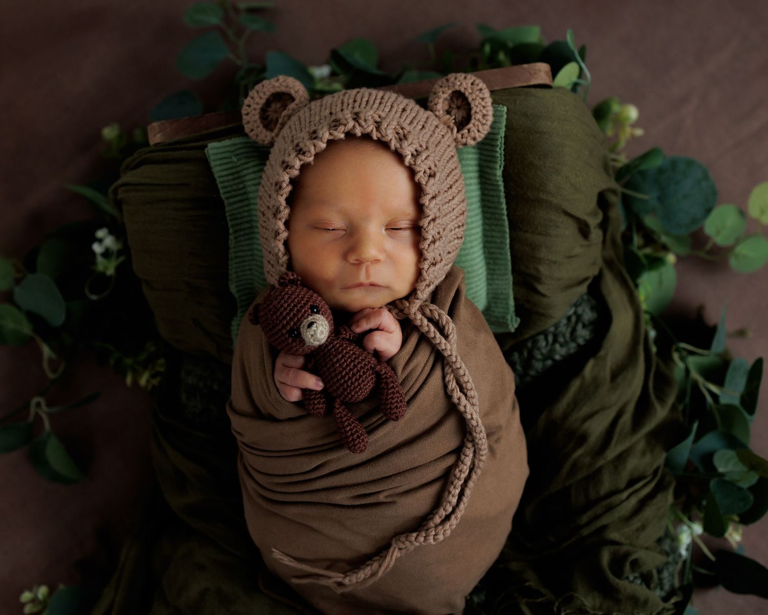 A sleeping newborn wrapped in a cozy brown bear-themed knit blanket lies surrounded by green foliage.