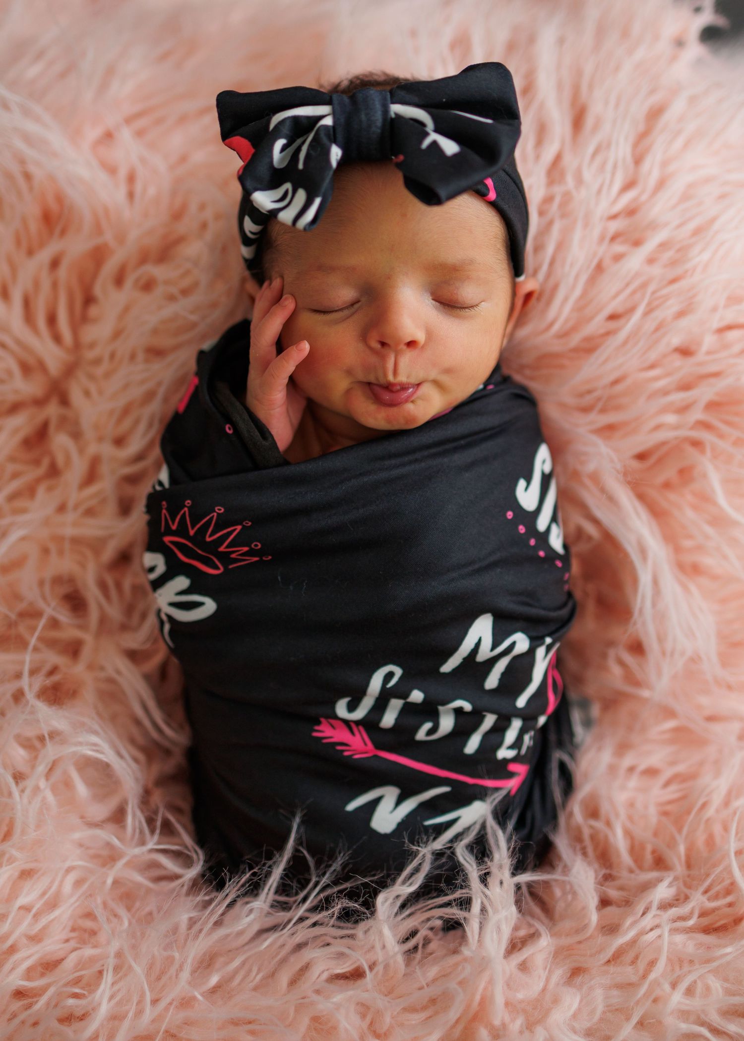 Sleeping newborn wrapped in black printed swaddle with bow headband resting on pink furry blanket.