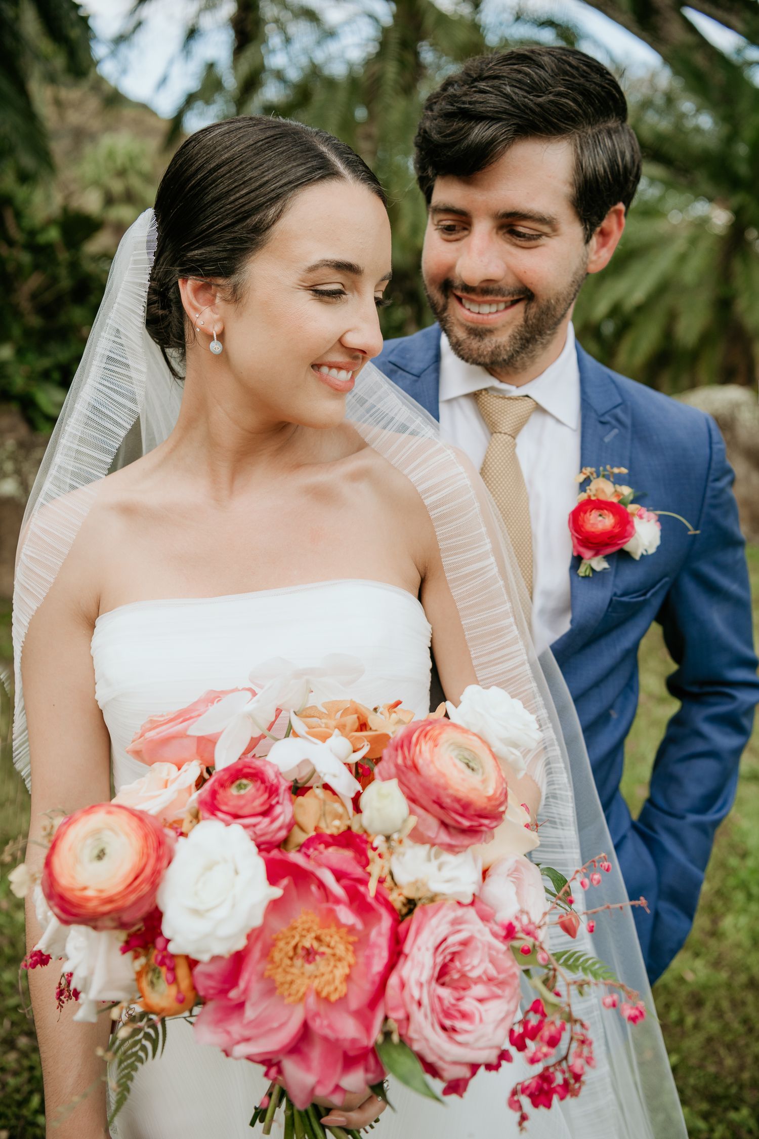 Wedding couple holding coral bouquet walk on grass with tropical palm trees and lush mountains in background.