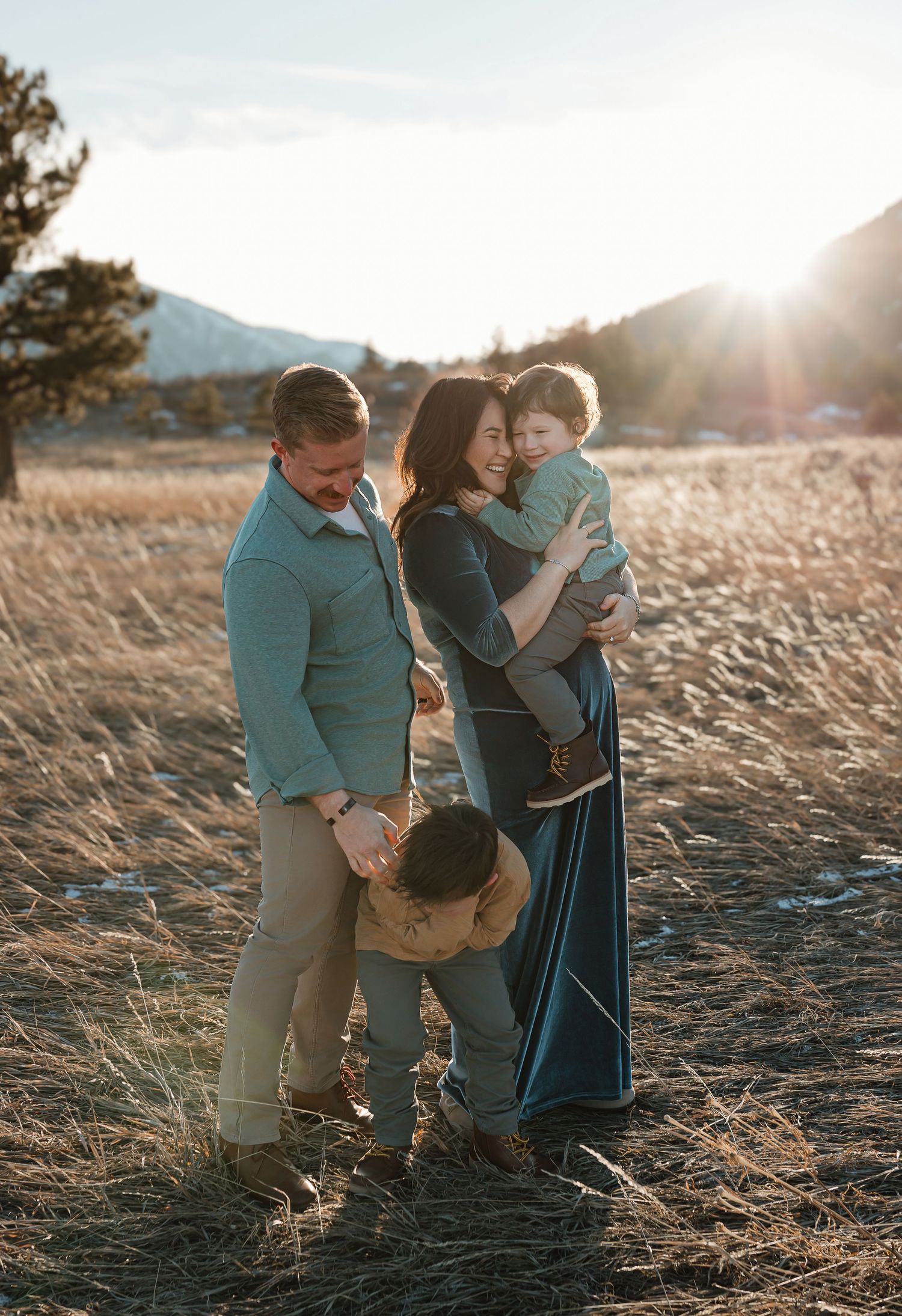 Couple with their child embrace in a golden field at sunset with mountains in the background.