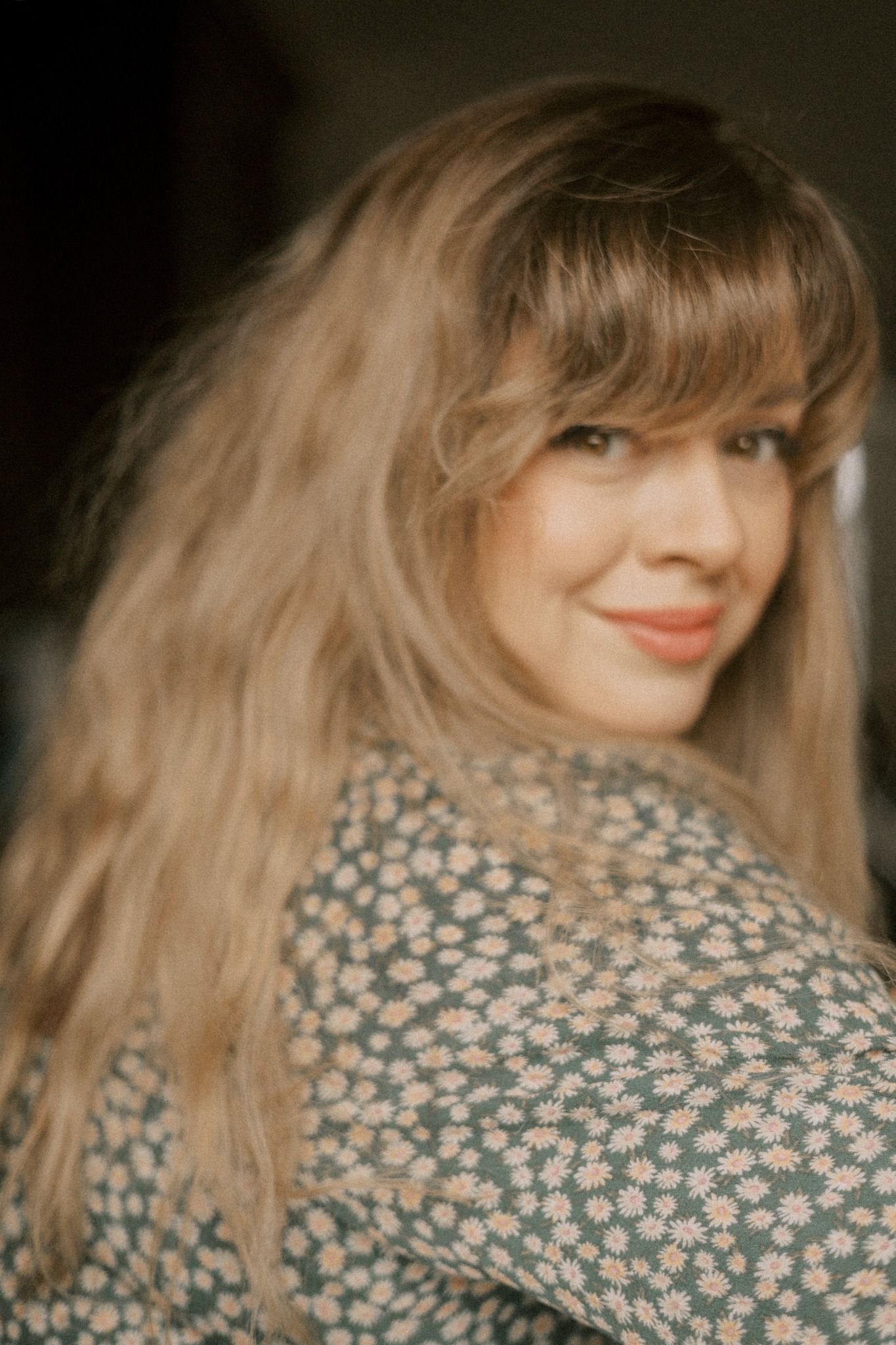 A woman in a floral print top smiles warmly while looking over her shoulder in a soft-focus portrait.