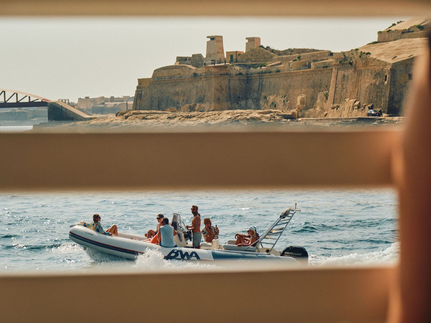 A group in an inflatable boat rides waves near coastal cliffs and a bridge at sunset.